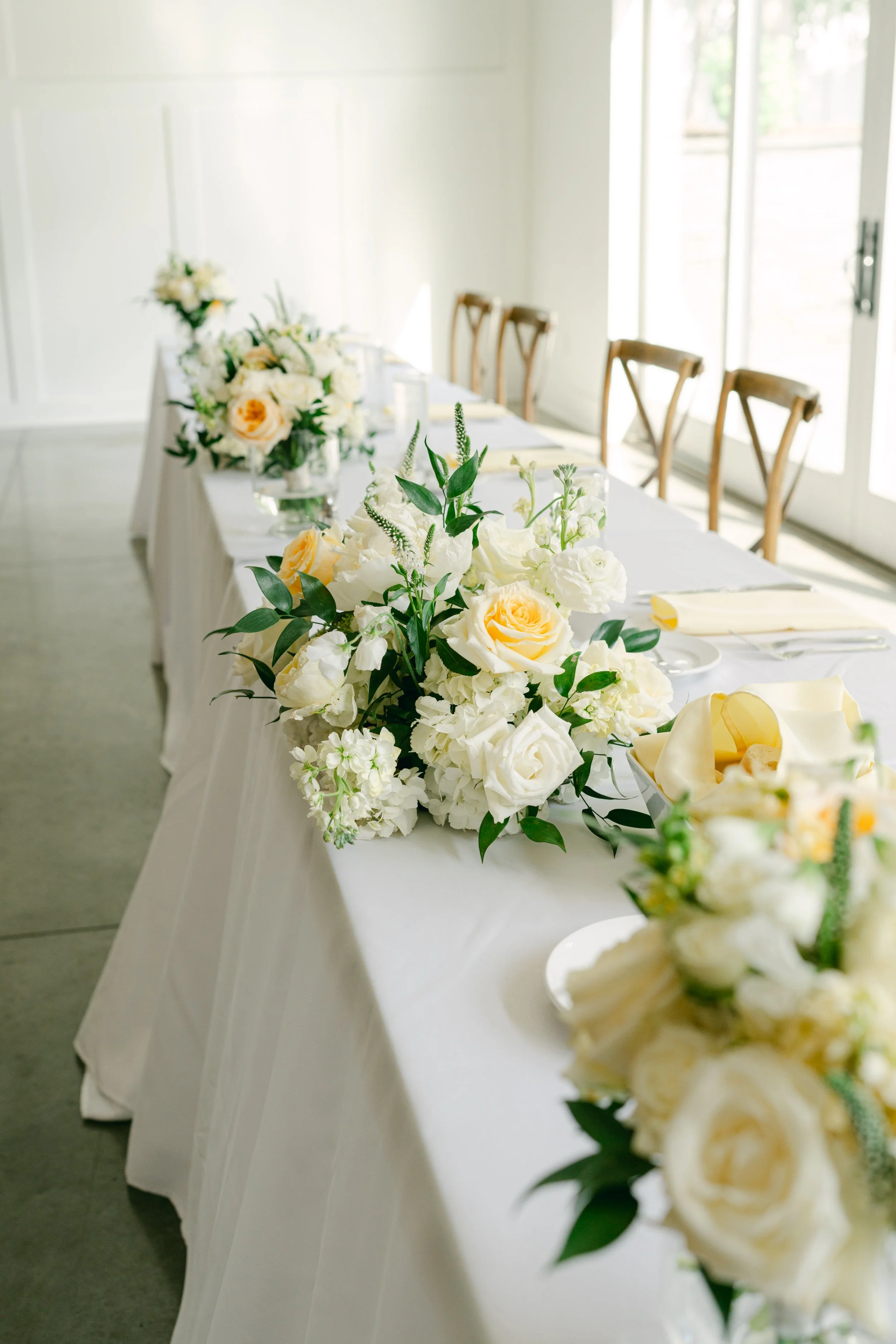 A long table decorated with white and yellow floral arrangements in a bright room with large windows.