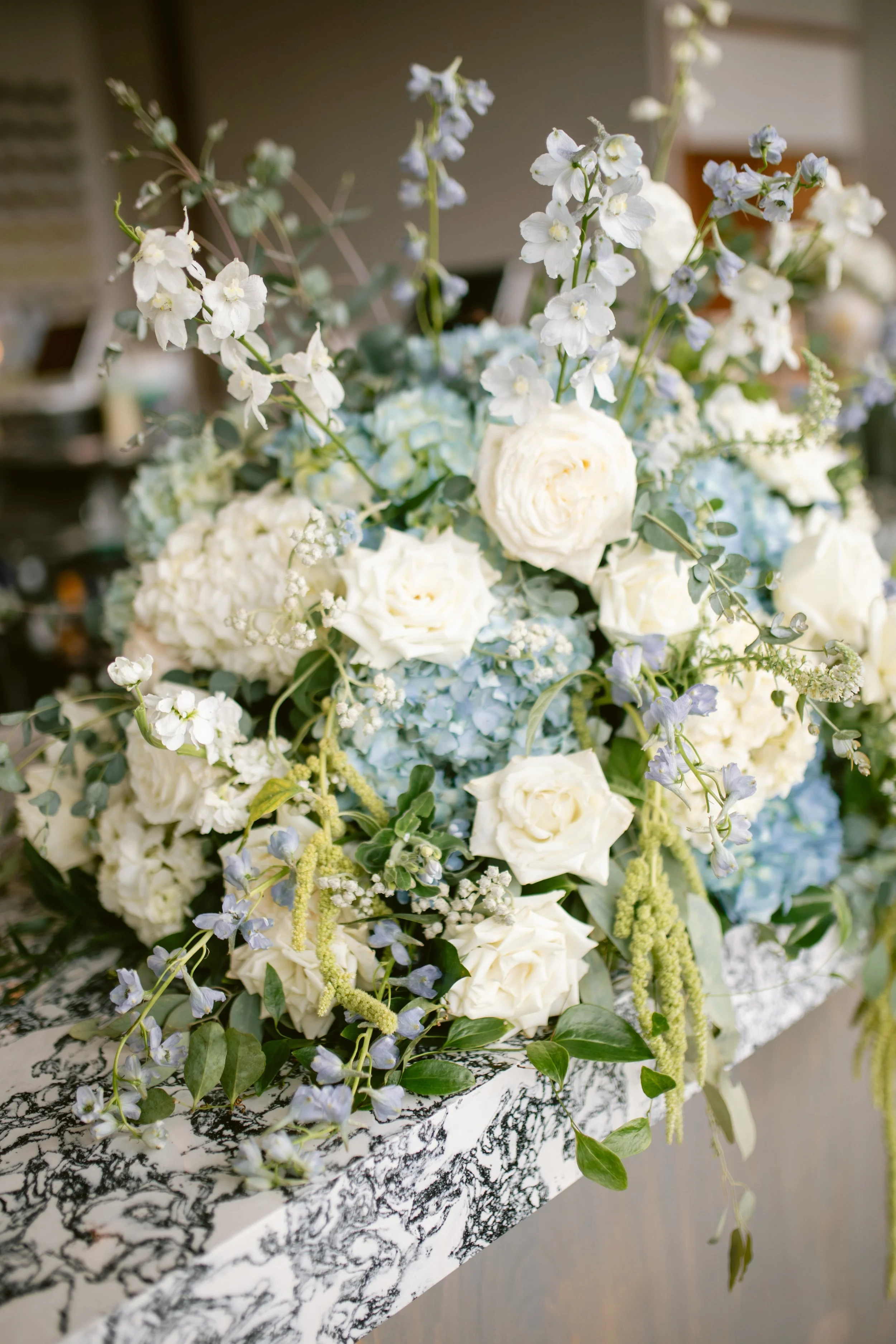 A floral arrangement with white roses, blue hydrangeas, and white delphiniums on a black-and-white patterned tablecloth.