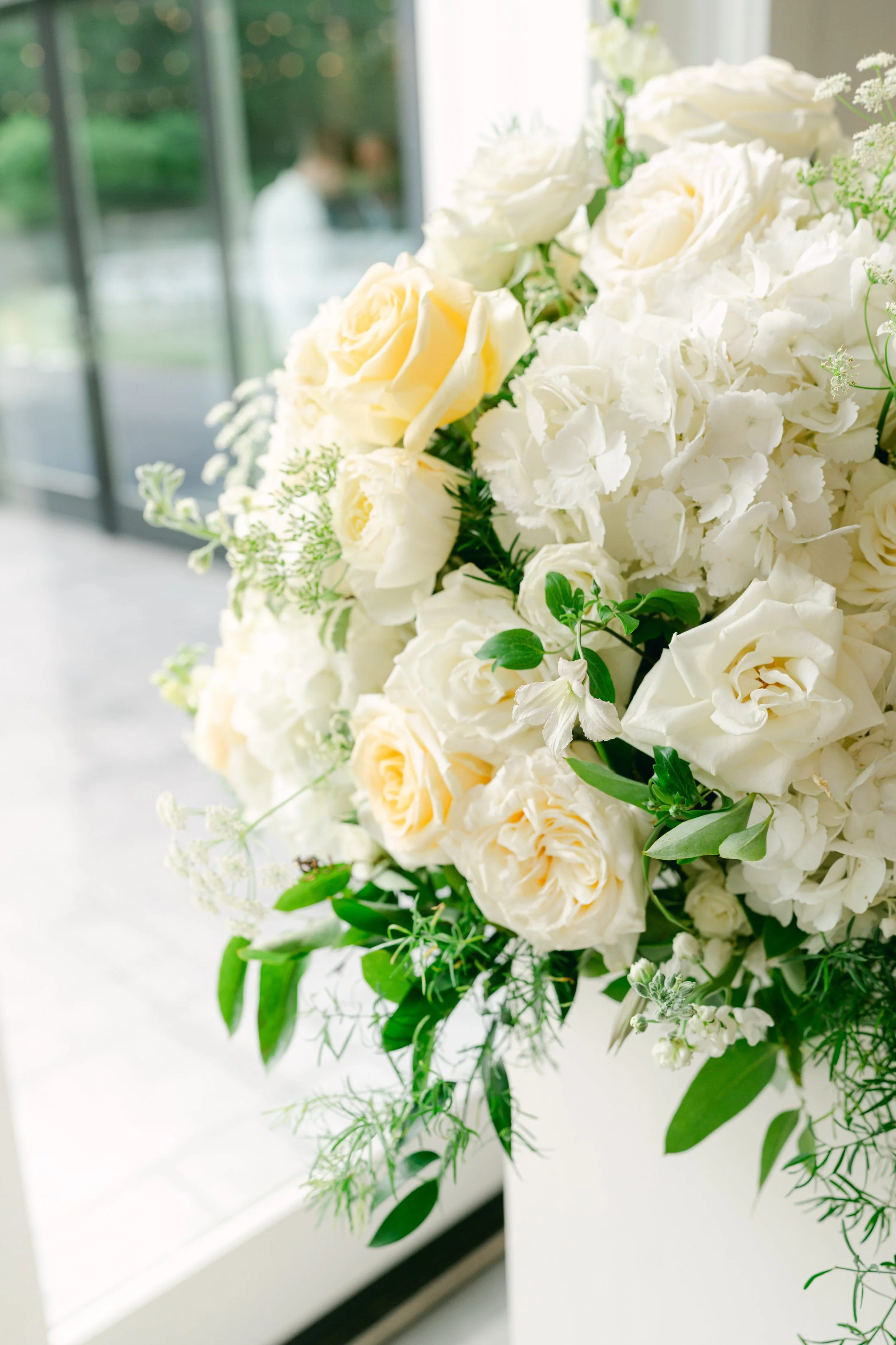 A large bouquet of white roses and greenery, with a blurred background of a window and a person inside a room.