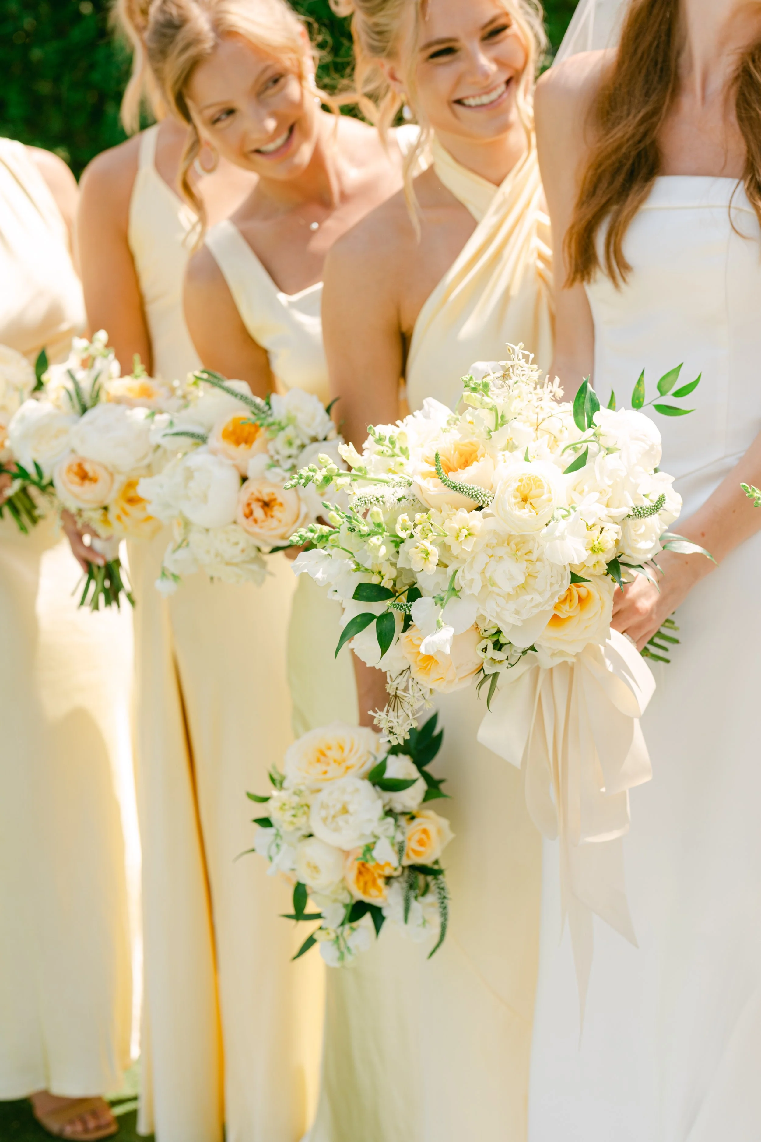 A line of women in yellow dresses holding white and peach floral bouquets at an outdoor wedding.