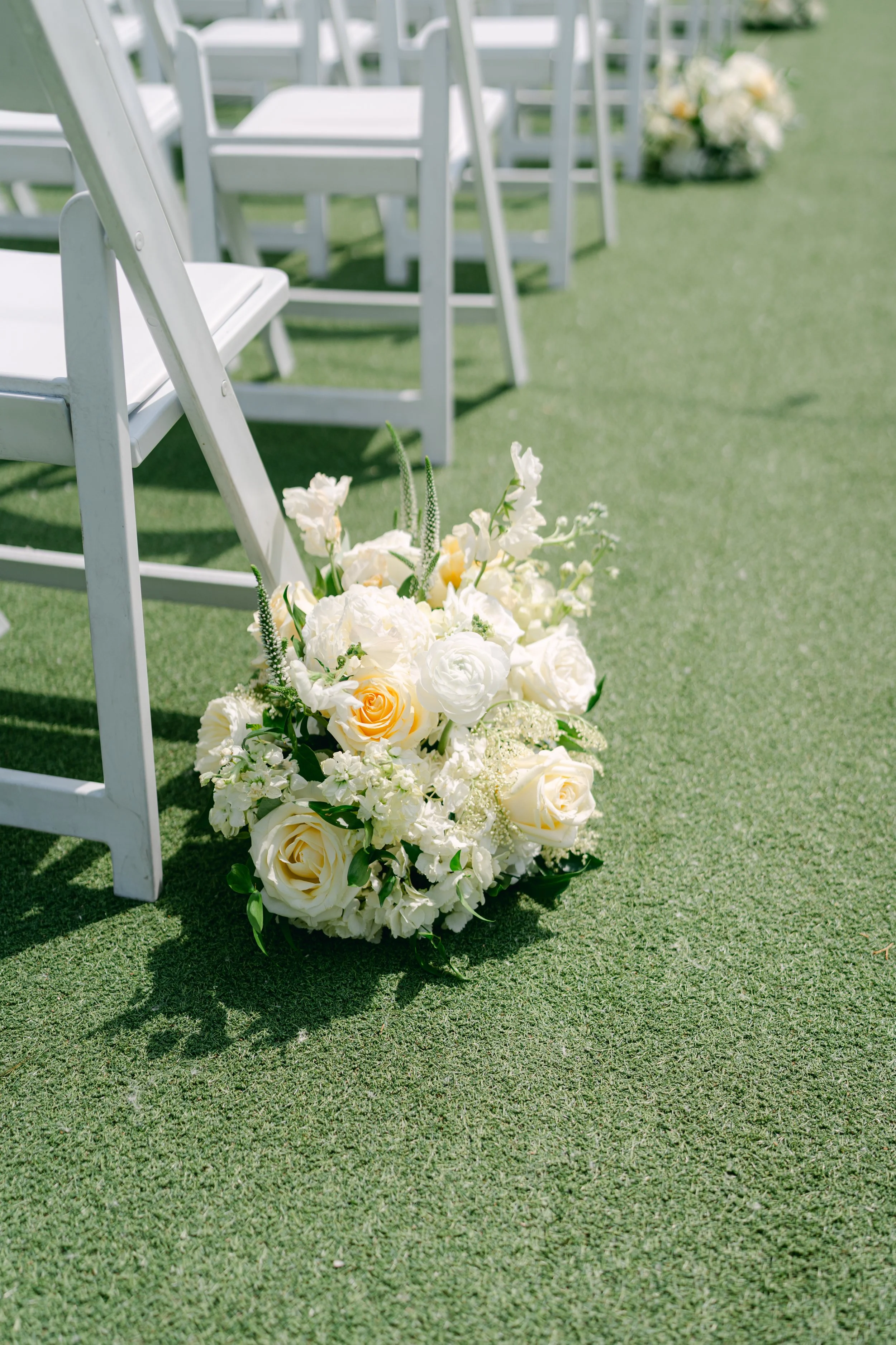 White chairs arranged on a grassy outdoor setting with a bouquet of white and cream flowers placed in front of the chairs, indicating a wedding ceremony setup.