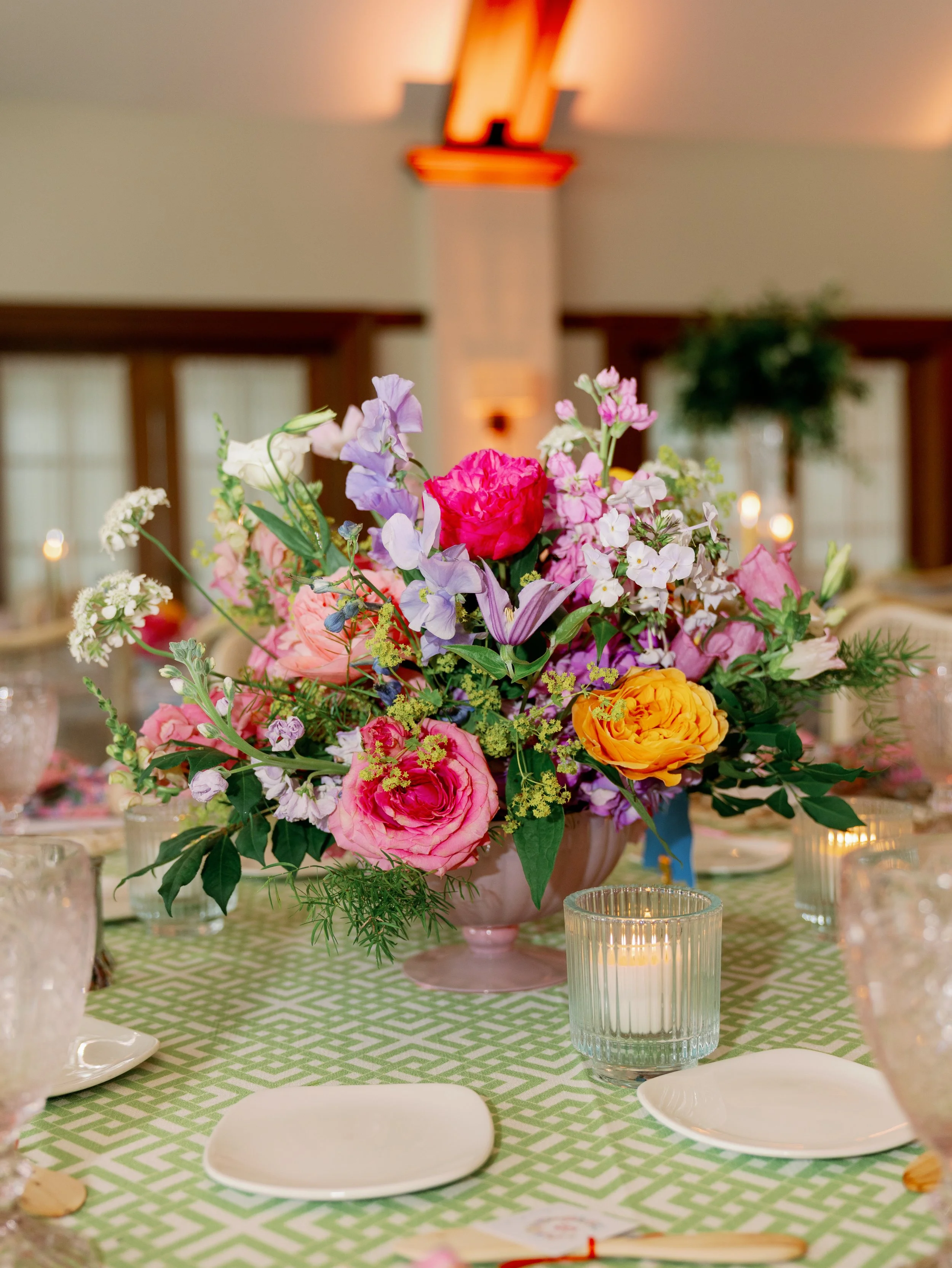 A colorful floral centerpiece with pink, purple, white, and orange flowers on a green and white patterned tablecloth, surrounded by white plates, clear vases, and small candles in glass holders in a decorated indoor setting.
