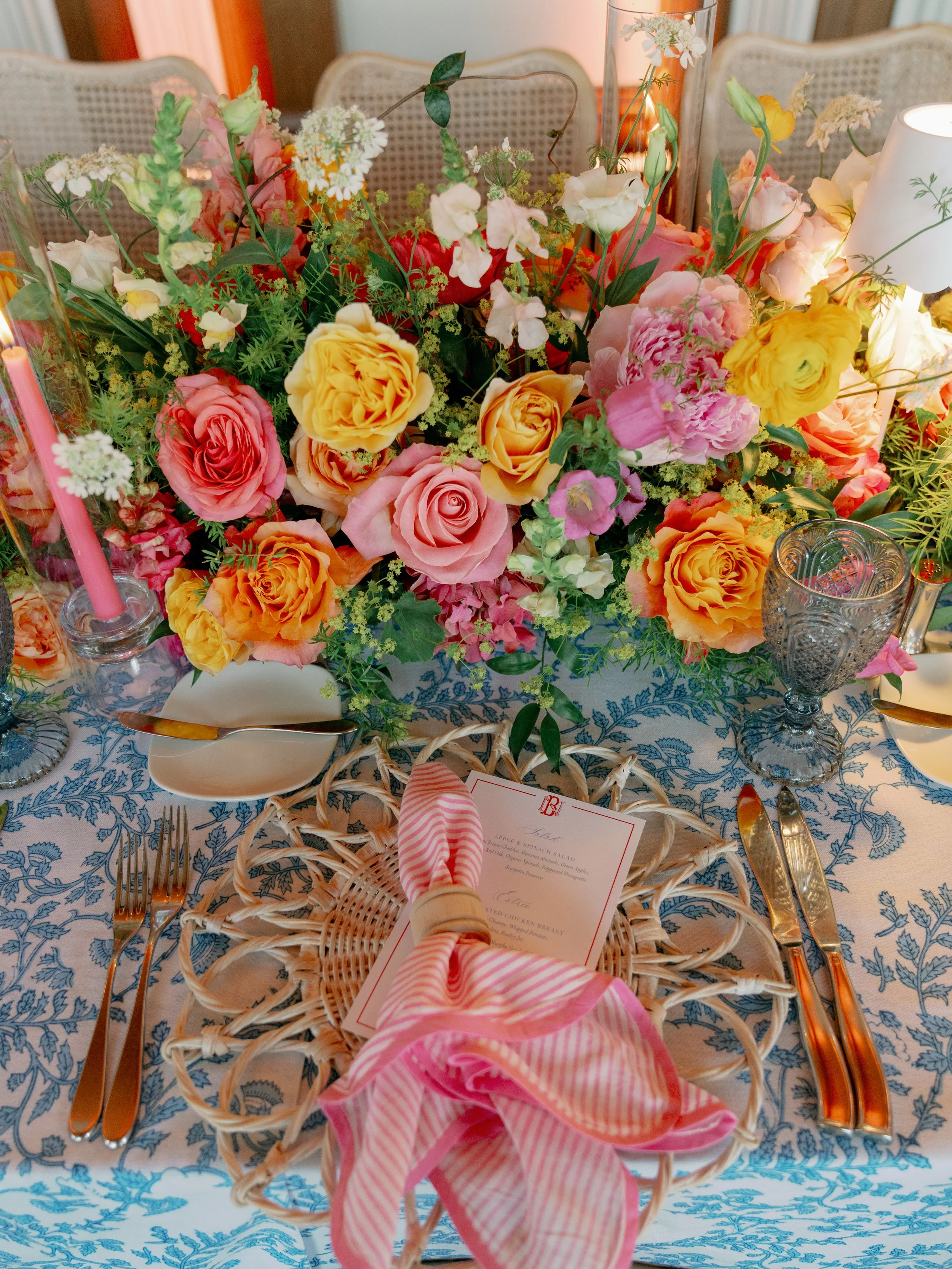 A formal dining table with a centerpiece of colorful flowers, including pink, yellow, white, and orange roses, set with glassware, silverware, and a pink and white striped napkin.