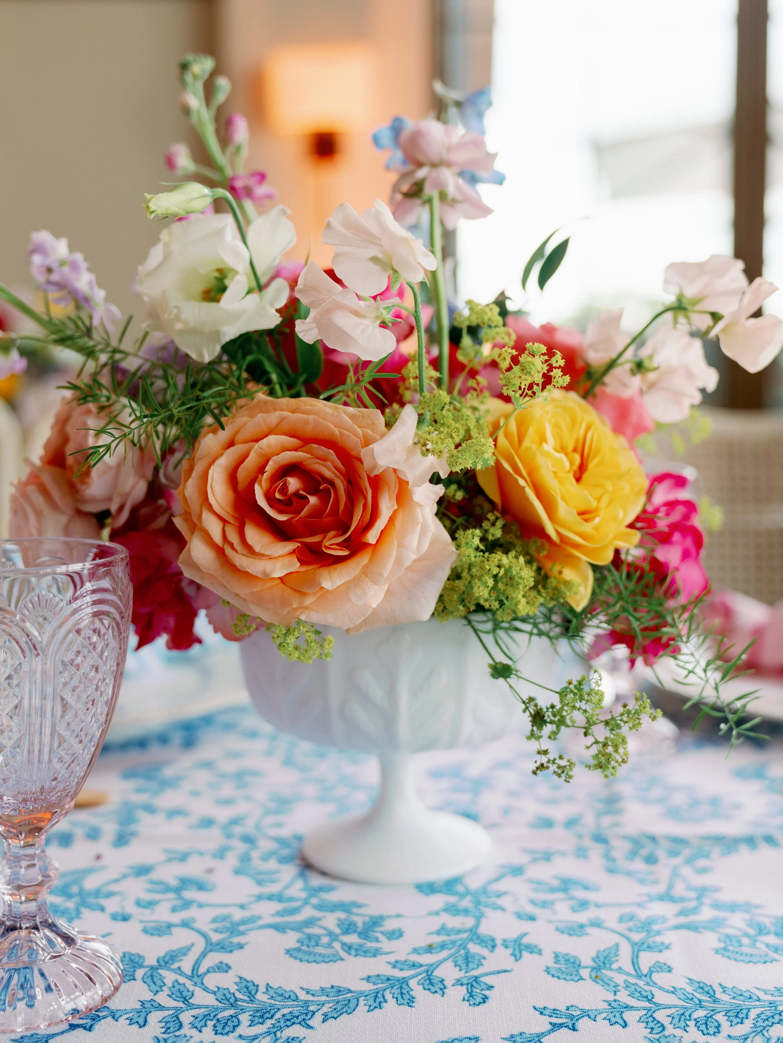 A floral centerpiece with peach roses, pink, white, and purple flowers, green leaves, and small green filler flowers on a table with a blue floral tablecloth. In the background, a blurred room with a lamp and window.