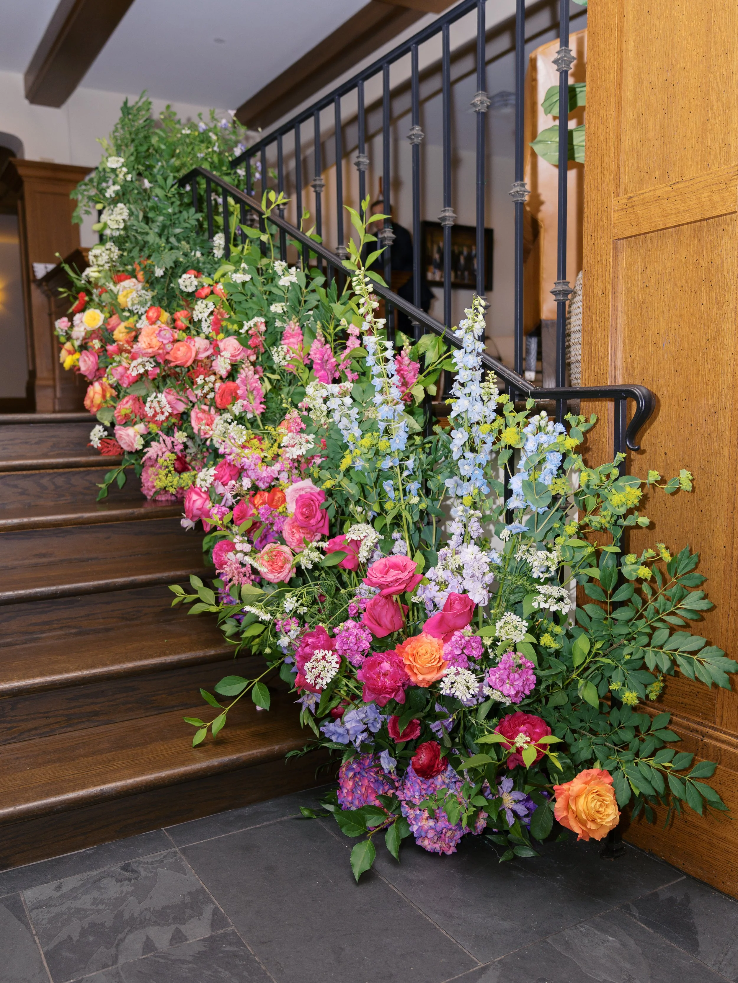 A large colorful floral arrangement is placed along the wooden stairs and railing in an indoor space, featuring pink, purple, yellow, orange, and white flowers with green foliage.