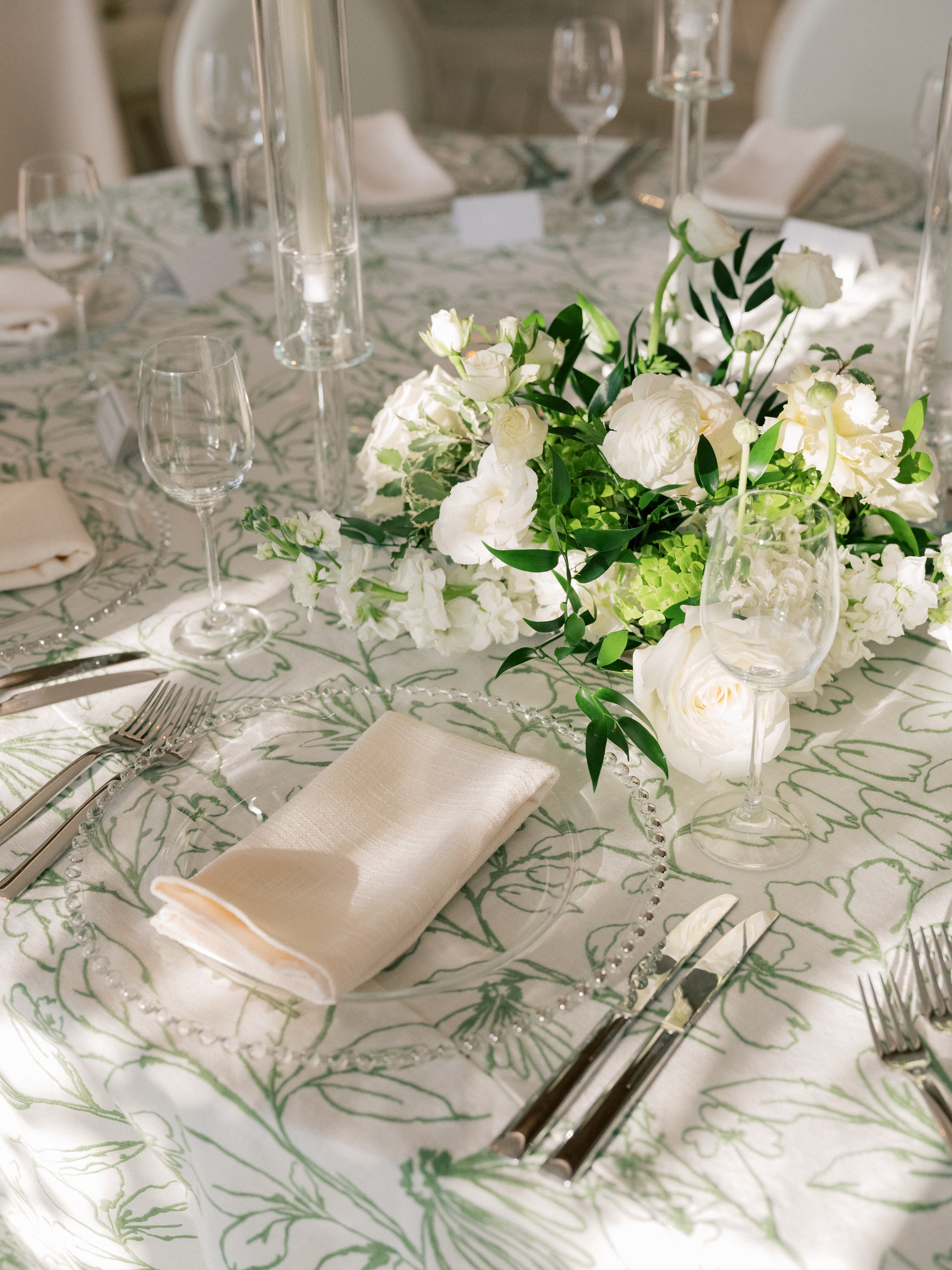 A table decorated with a white and green floral centerpiece, set with glassware, silverware, a white folded napkin, and a patterned tablecloth for a formal event.