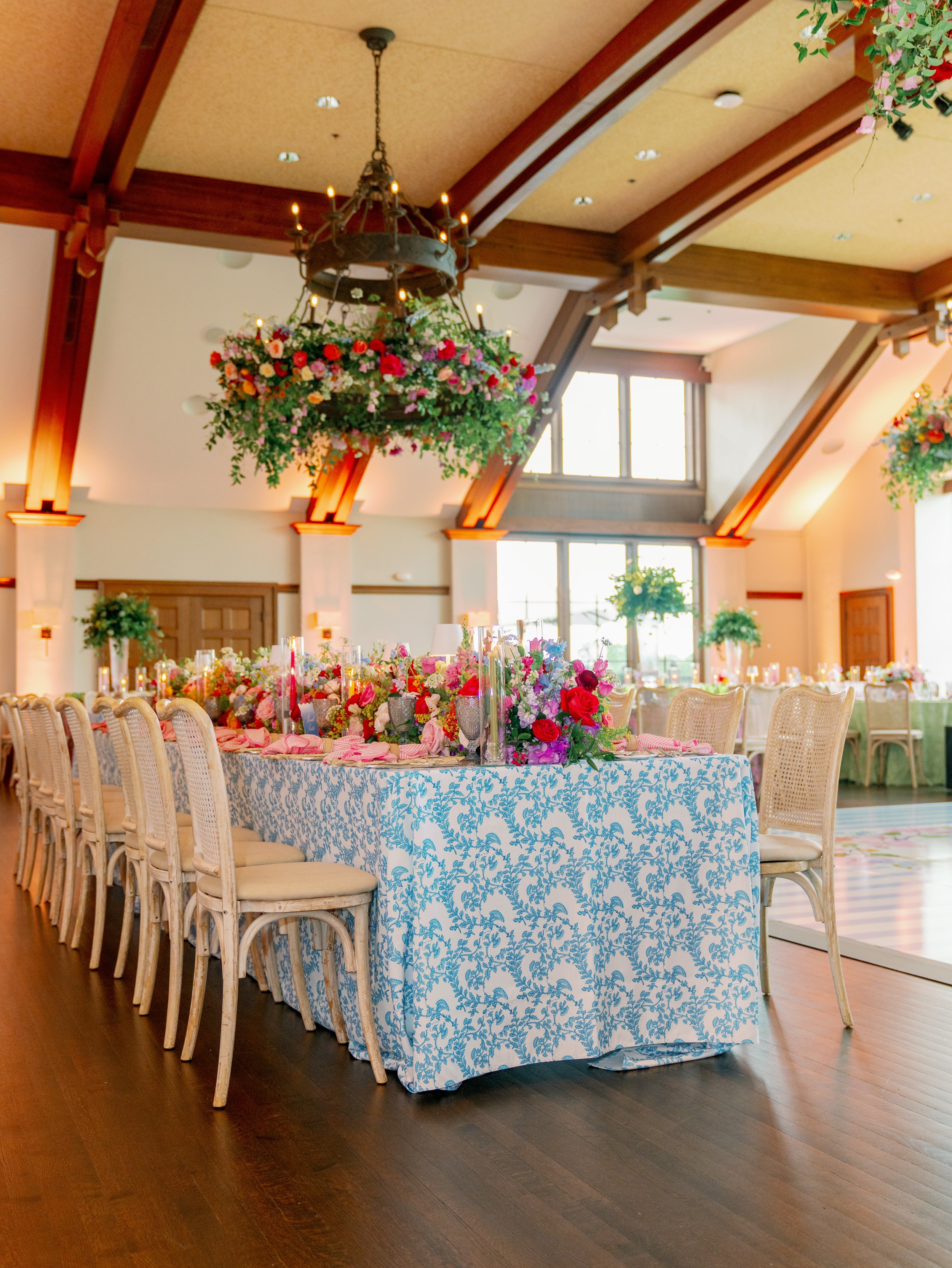 Indoor event space decorated with floral arrangements on tables and hanging floral chandelier, with large windows and wooden ceiling beams.