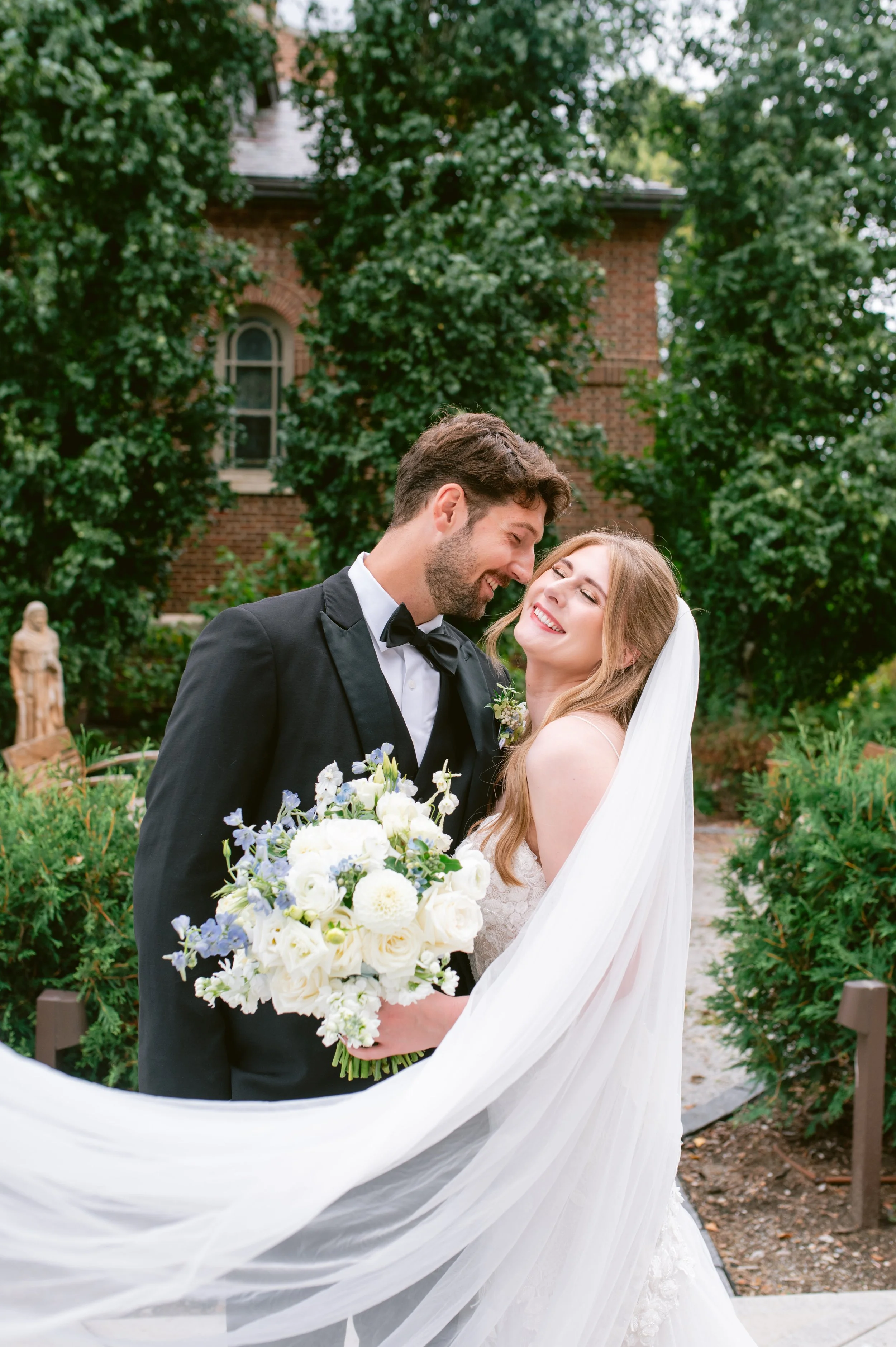 A bride and groom dressed in formal wedding attire sharing a joyful moment outdoors, surrounded by greenery and a brick building in the background.
