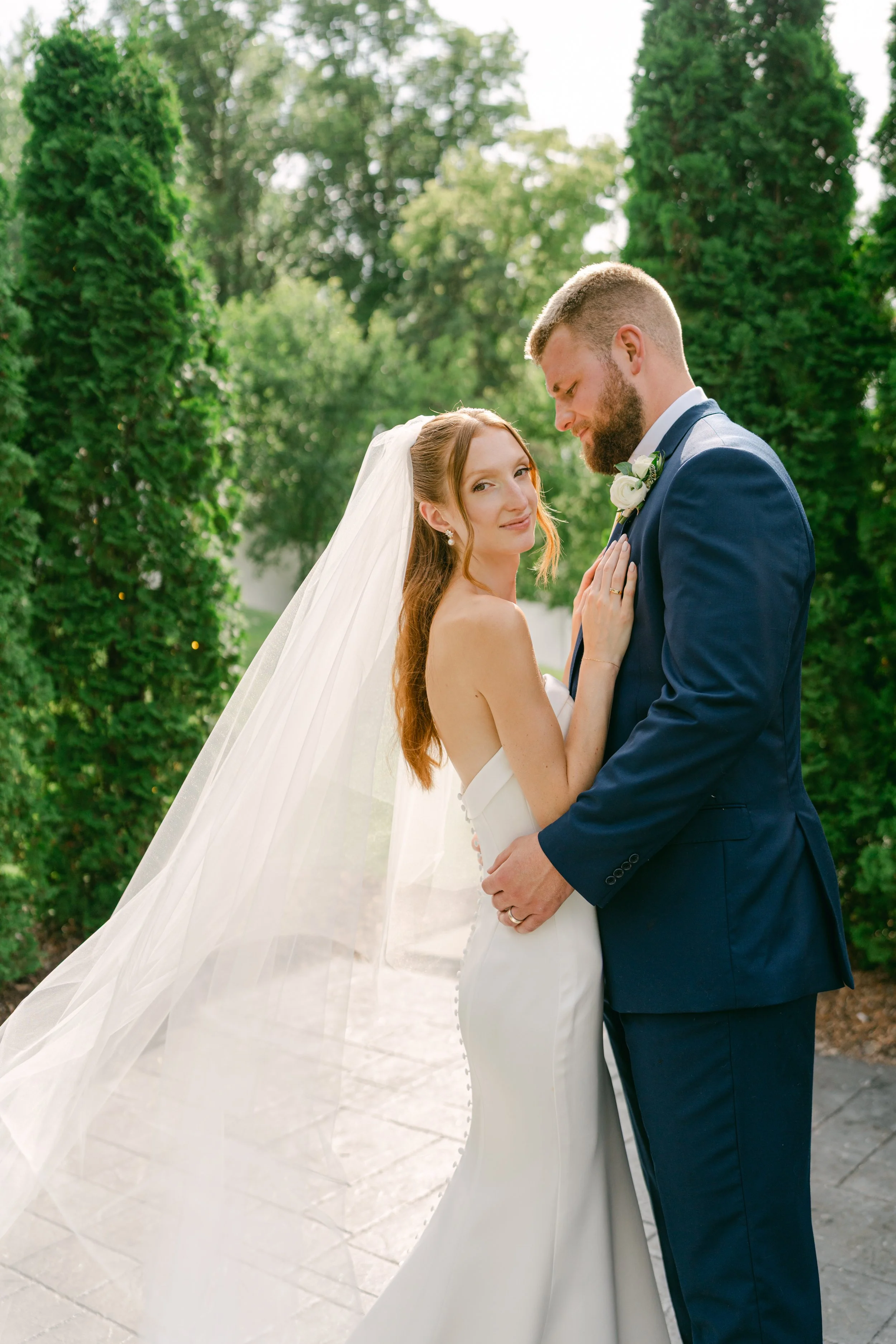 A bride and groom standing close together outdoors, surrounded by tall green trees and a bright sky in the background. The bride is wearing a white wedding dress and veil, while the groom is dressed in a blue suit with a boutonniere. They are gazing 