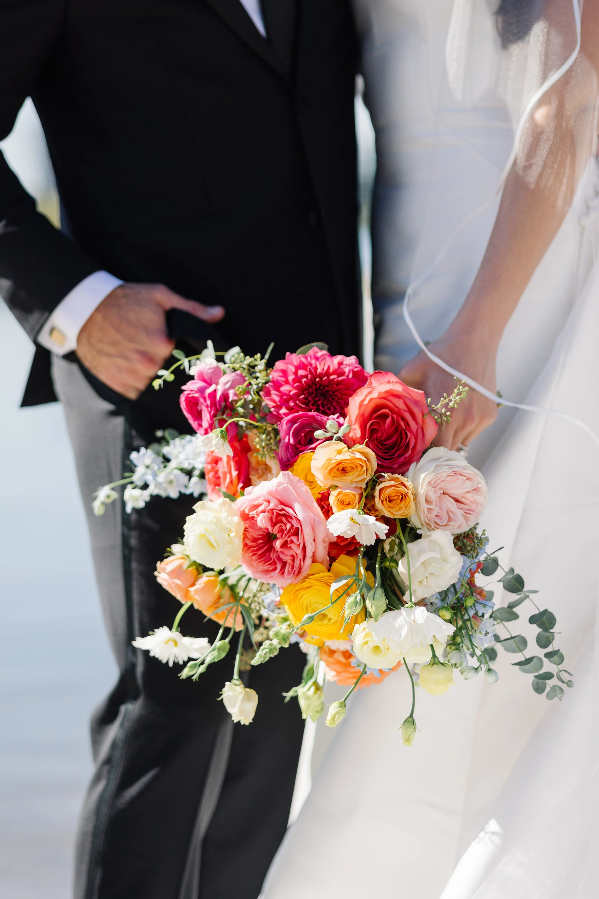 Close-up of a bride and groom holding a colorful bouquet of flowers during a wedding.