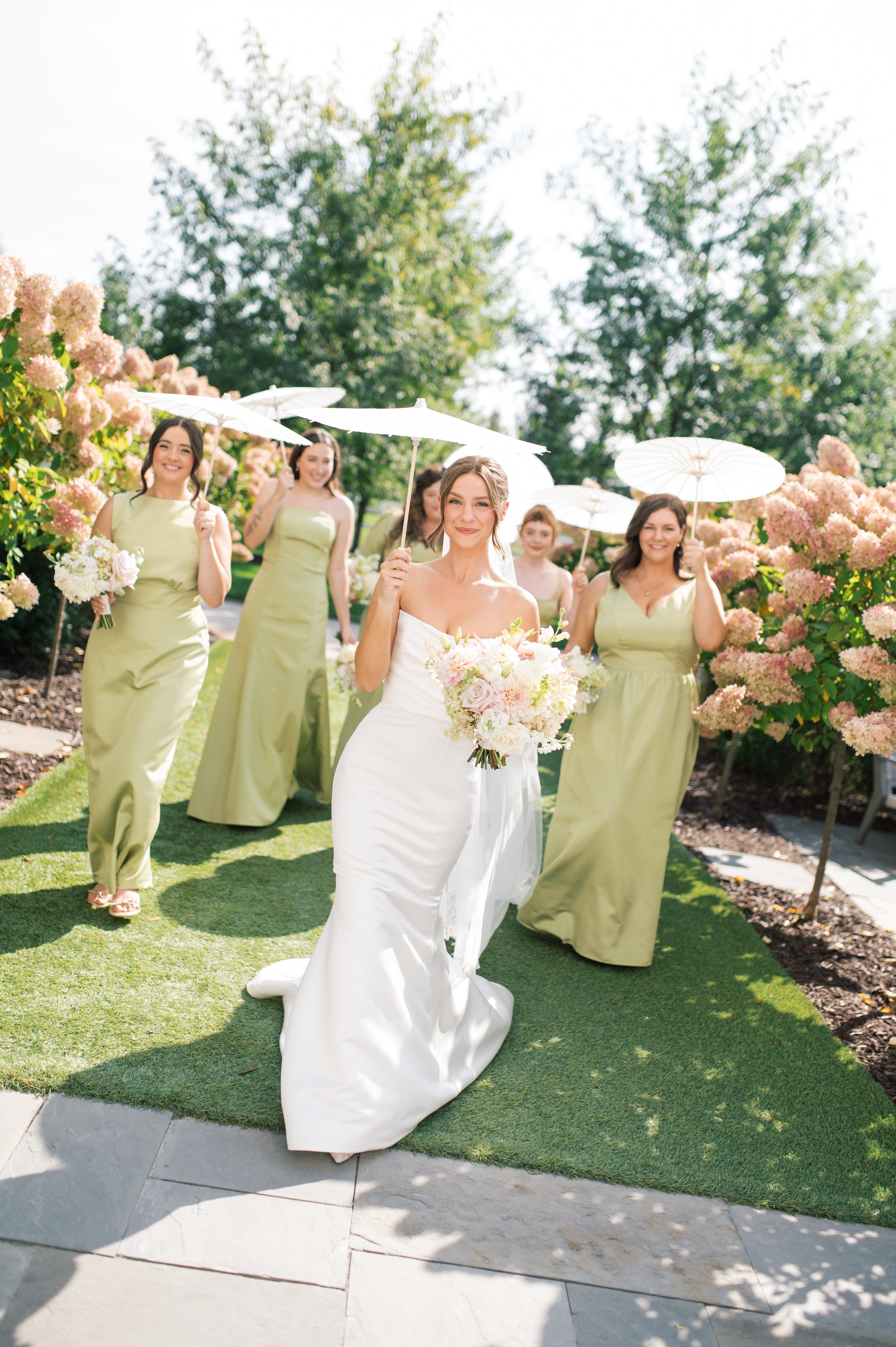 A bride in a white wedding dress holding a bouquet, walking outdoors with five bridesmaids in light green dresses, holding white umbrellas, surrounded by pink flowers and green trees.