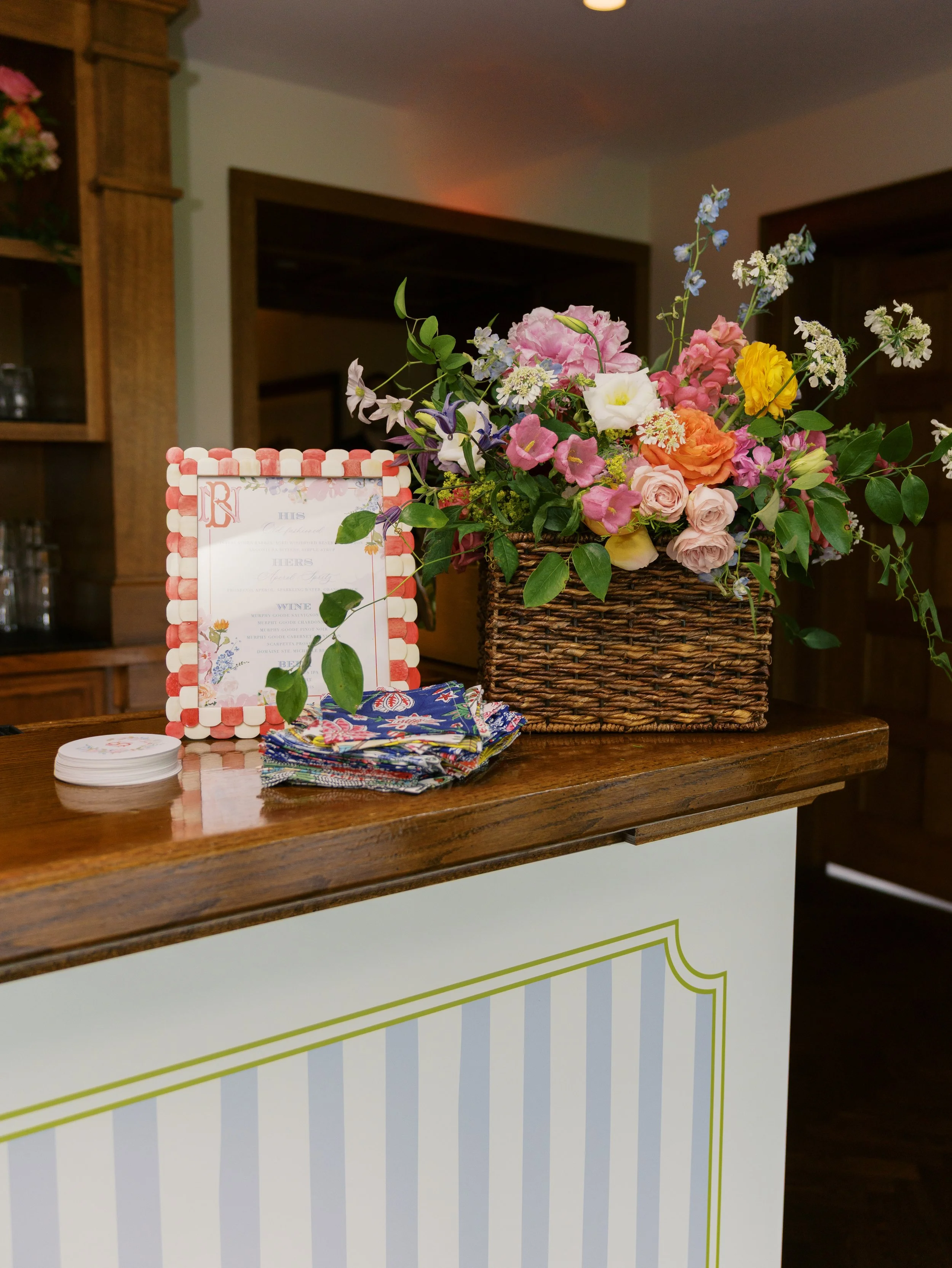 A floral arrangement in a wicker basket on a wooden table, with a decorative frame and a menu or sign nearby, in a cozy indoor setting.