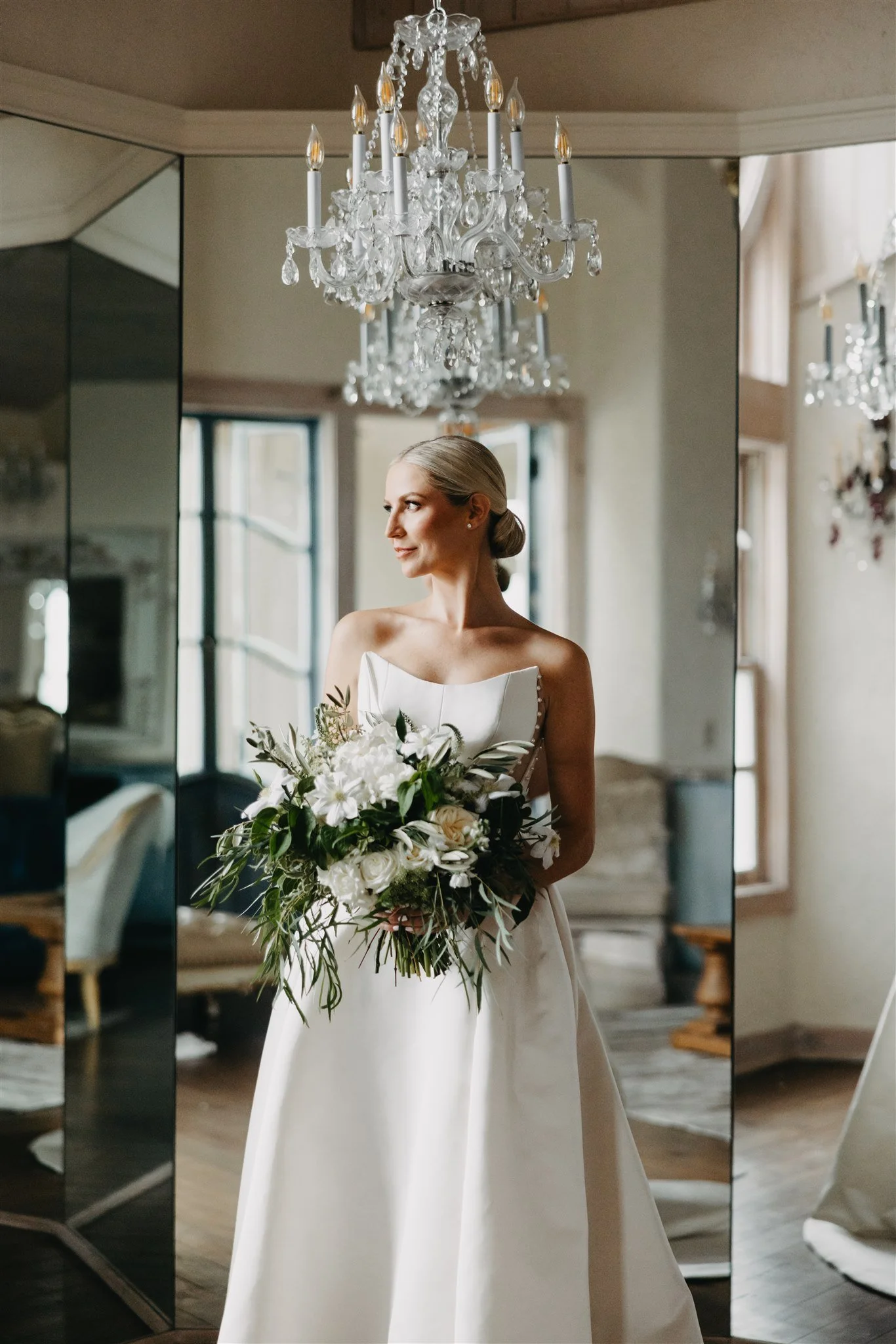 A bride in a white wedding gown holding a large bouquet of white flowers and greenery standing in a room with a chandelier and mirrors.