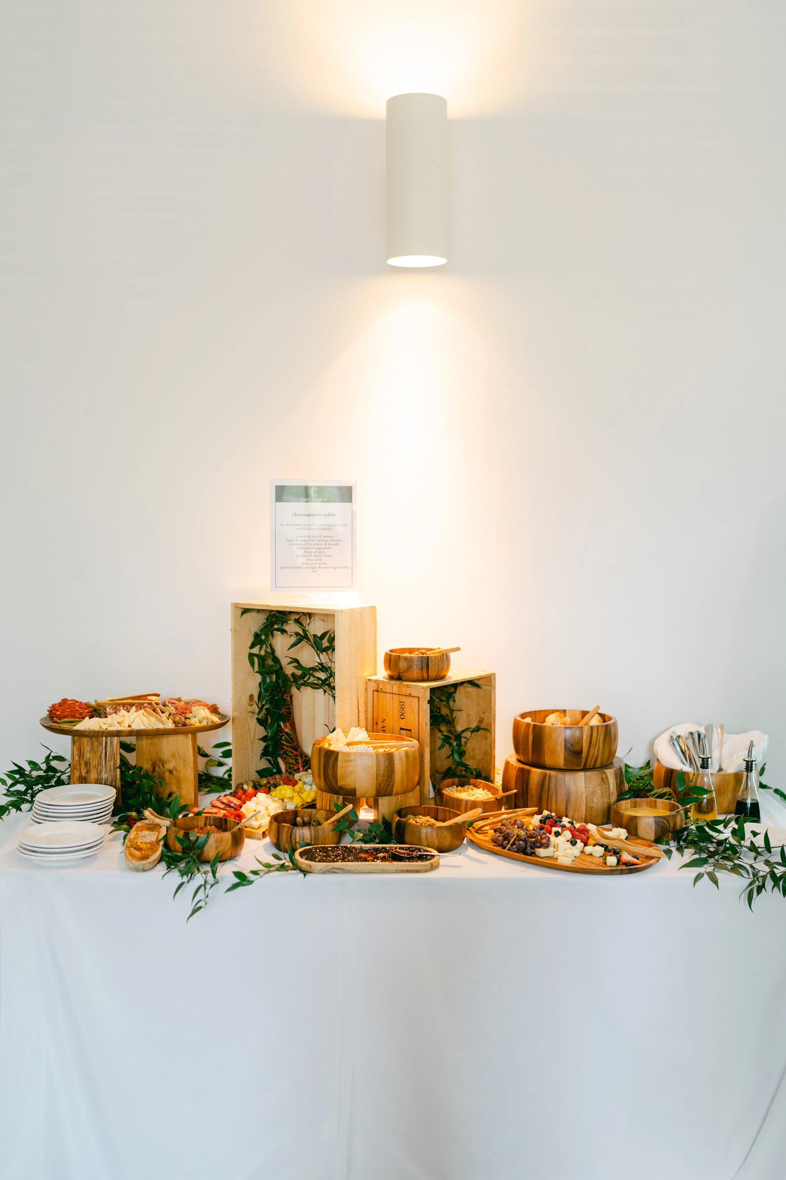Buffet table with assorted cheeses, fruits, meats, and crackers, arranged on wooden bowls and platters, decorated with green leaves.