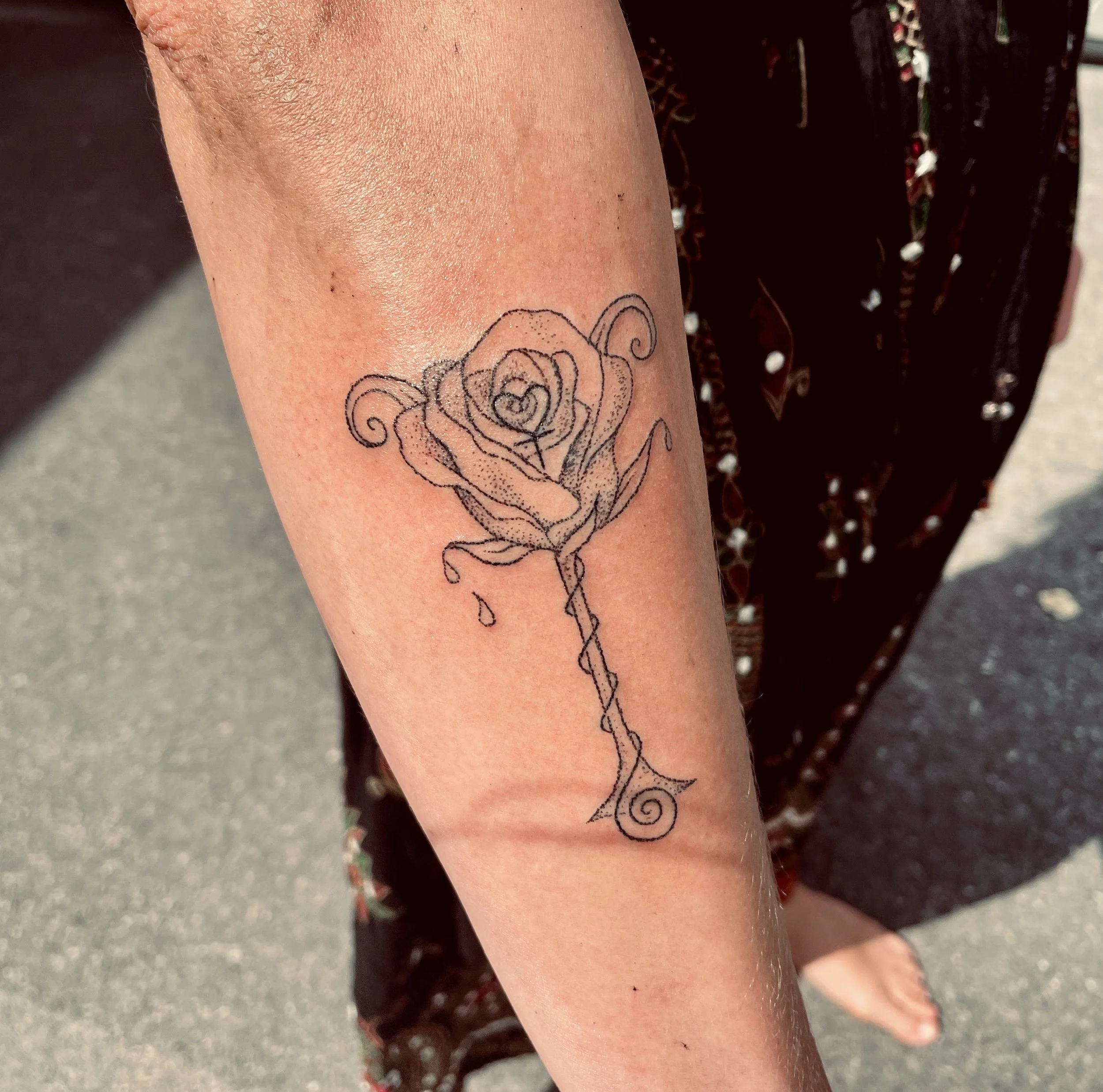 Close-up of a person's forearm with a tattoo of a stylized rose and leaves, featuring heart shapes within the floral design.