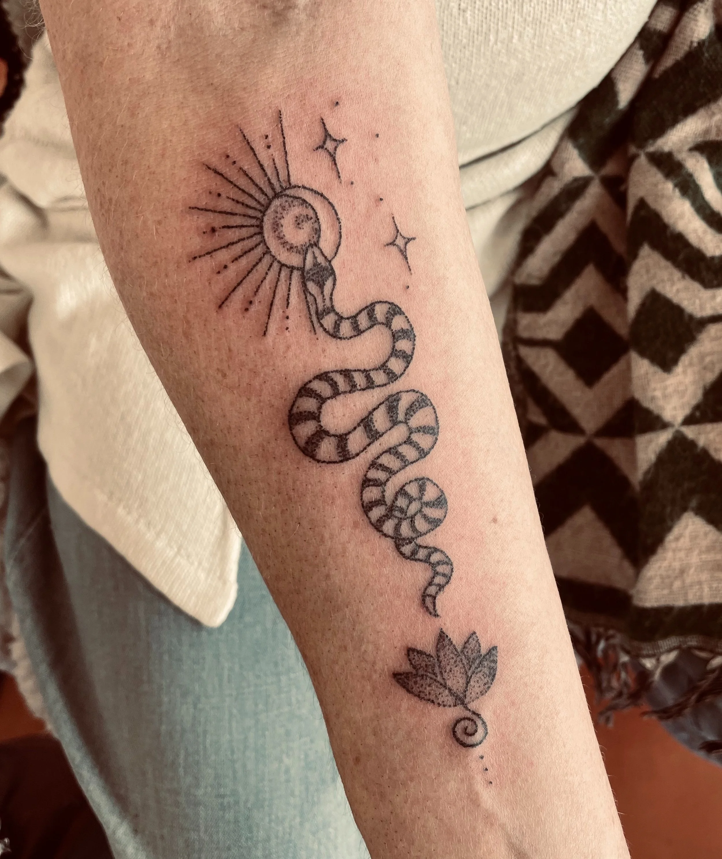 Black line tattoo of a zodiac snake wrapped around a celestial sun with rays, stars, a leaf, and decorative elements on a person's arm.