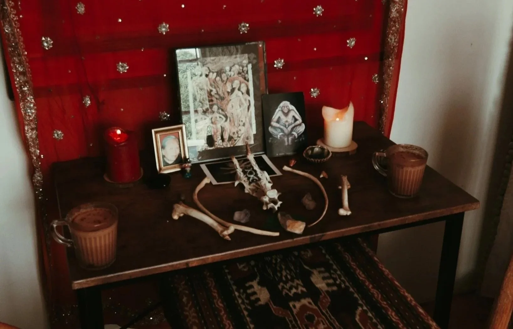 Tattoo Ceremony Altar with photos, candles, crystals, skull, and drink on a table with a red and gold decorated back panel