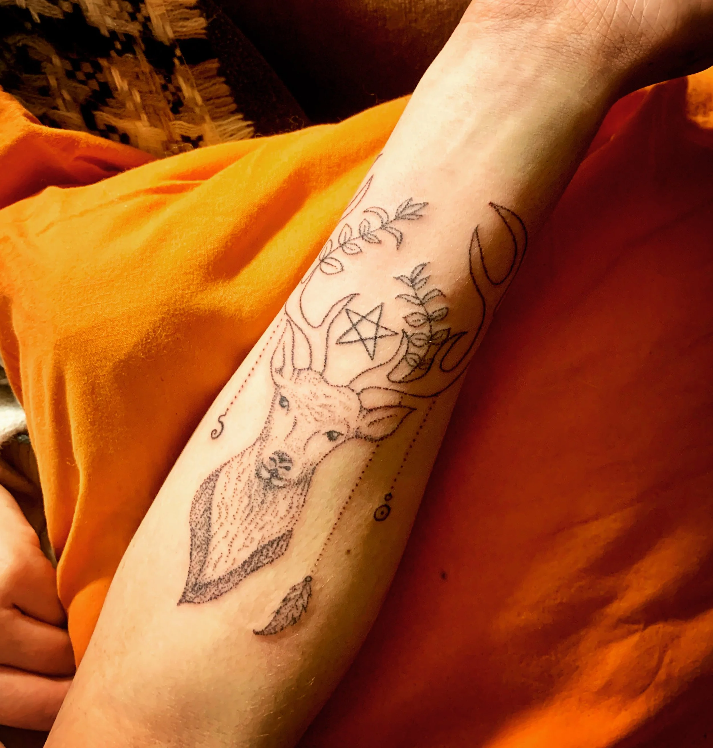Close-up of a person's arm with a skeleton and moon tattoo, wrapped in an orange blanket, with a woven fabric background.