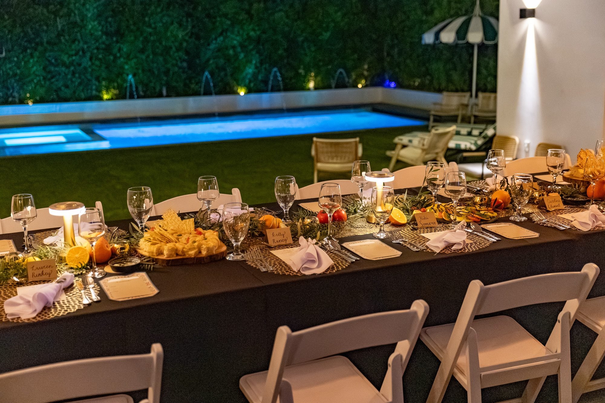 Long dining table set for a private event beside the lit backyard pool at night