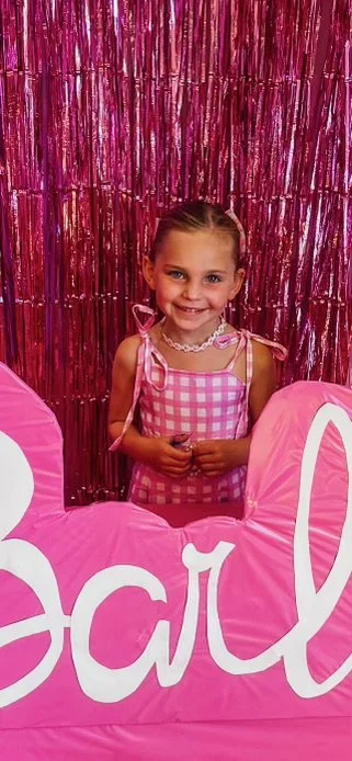 A young girl smiling in front of a shiny pink backdrop, holding a large pink sign with white cursive writing.