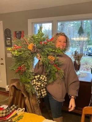 Woman holding a Christmas wreath decorated with pinecones, ribbons, and ornaments in a cozy home interior.