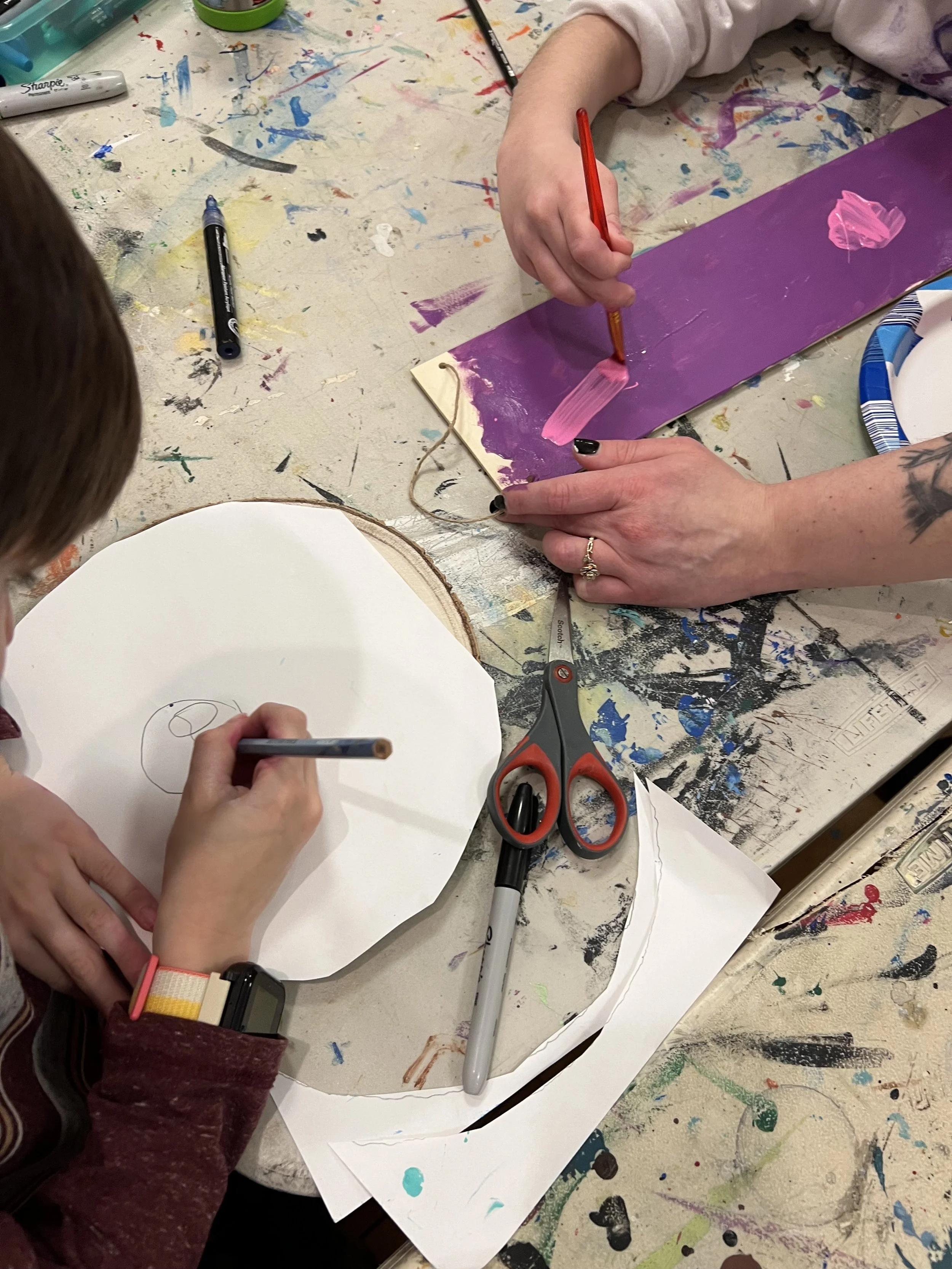 Child and adult working on a craft project at a messy table with paint and scissors, creating a painted design on a purple piece of paper.