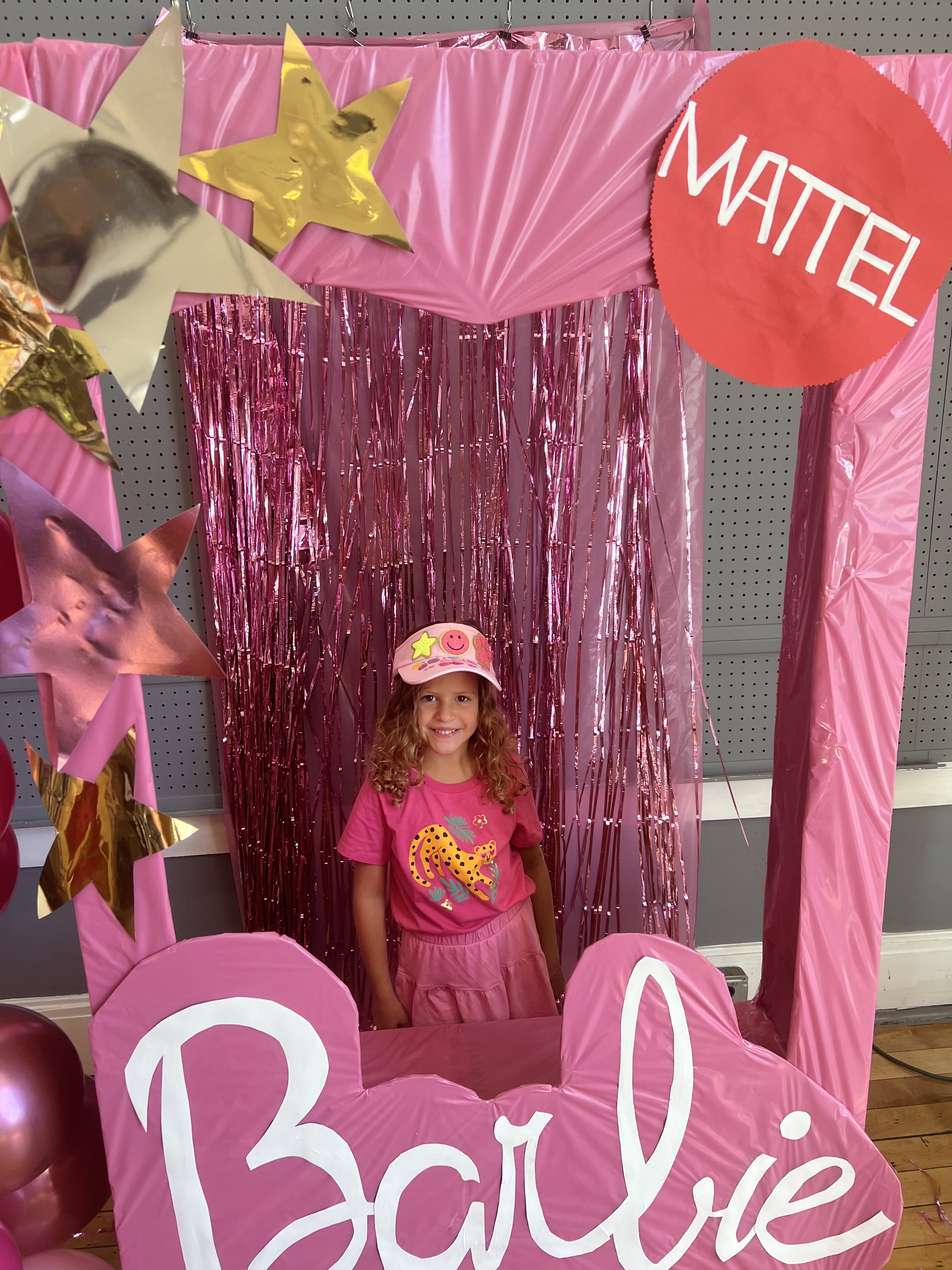 A young girl with curly hair smiling, wearing a pink cap and pink outfit with a dinosaur print, standing behind a pink photo booth decorated for a Barbie-themed event. The backdrop features pink metallic streamers, and the booth is labeled "Barbie" i