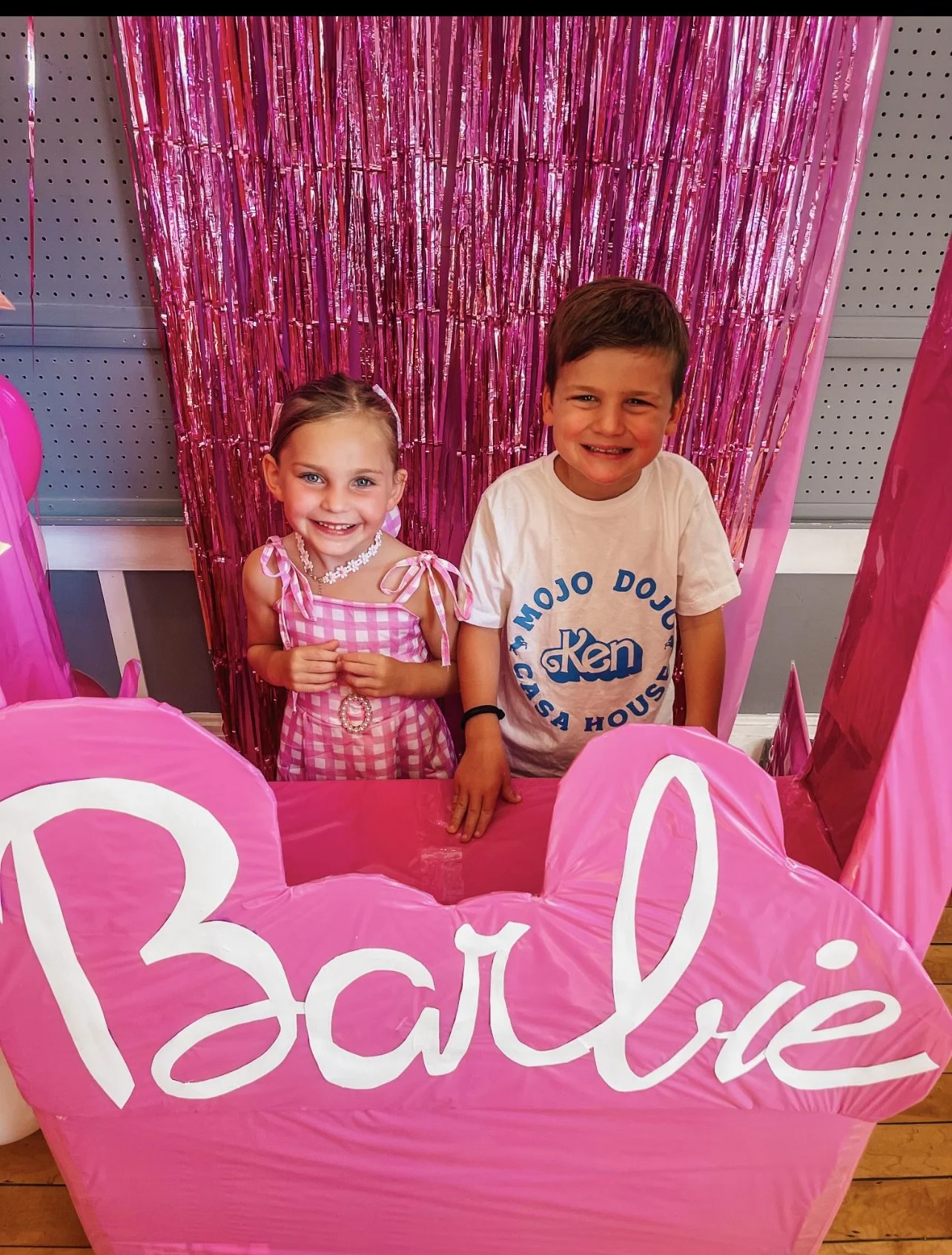 Two children, a girl and a boy, smiling behind a pink decorated area with a large pink heart-shaped sign that says 'Babies' in white letters. The girl is wearing a pink gingham dress with bows on the shoulders, and the boy is wearing a white t-shirt.