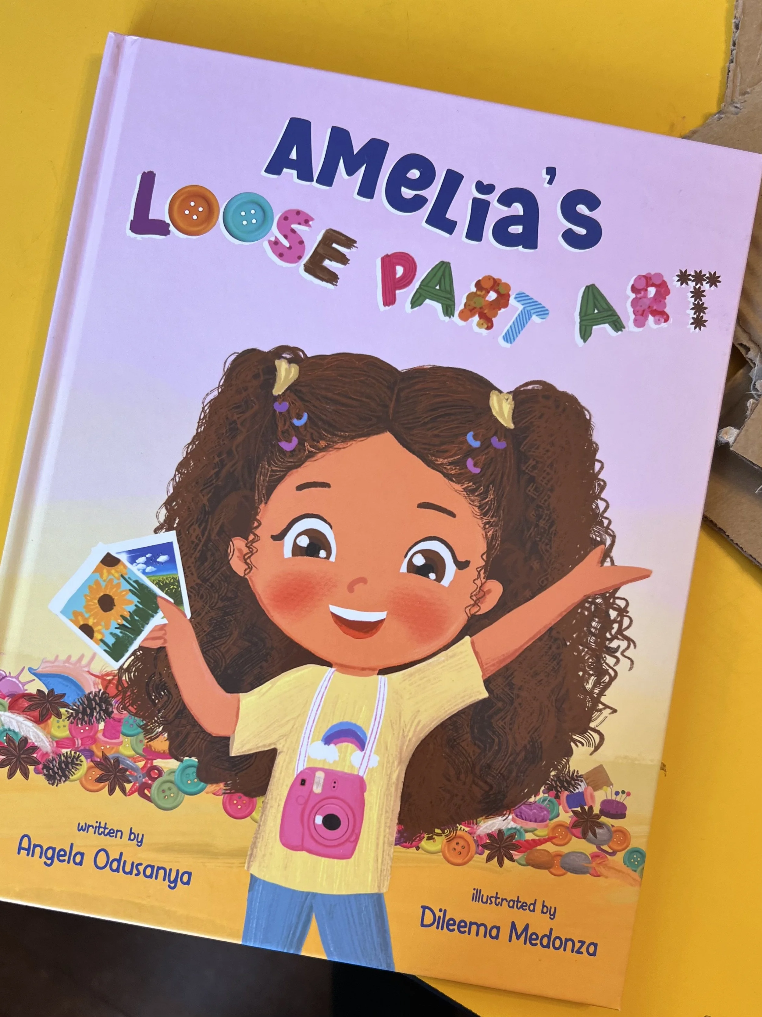 Children's book titled 'Amelia's Loose Papers' with an illustration of a happy girl with curly hair holding photographs, surrounded by buttons and colorful decorations.