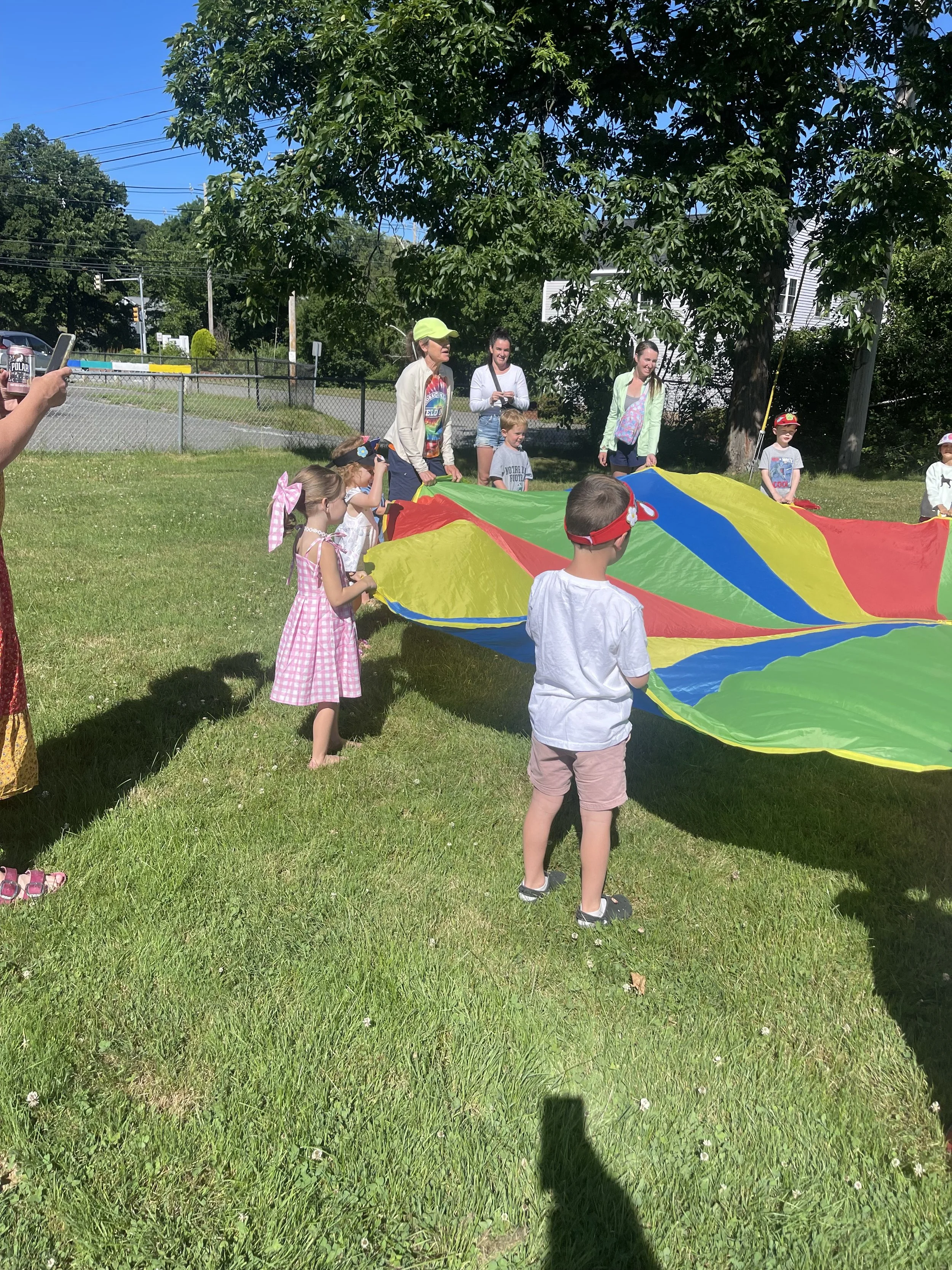 Children with adults playing with a colorful parachute outdoors on a sunny day.
