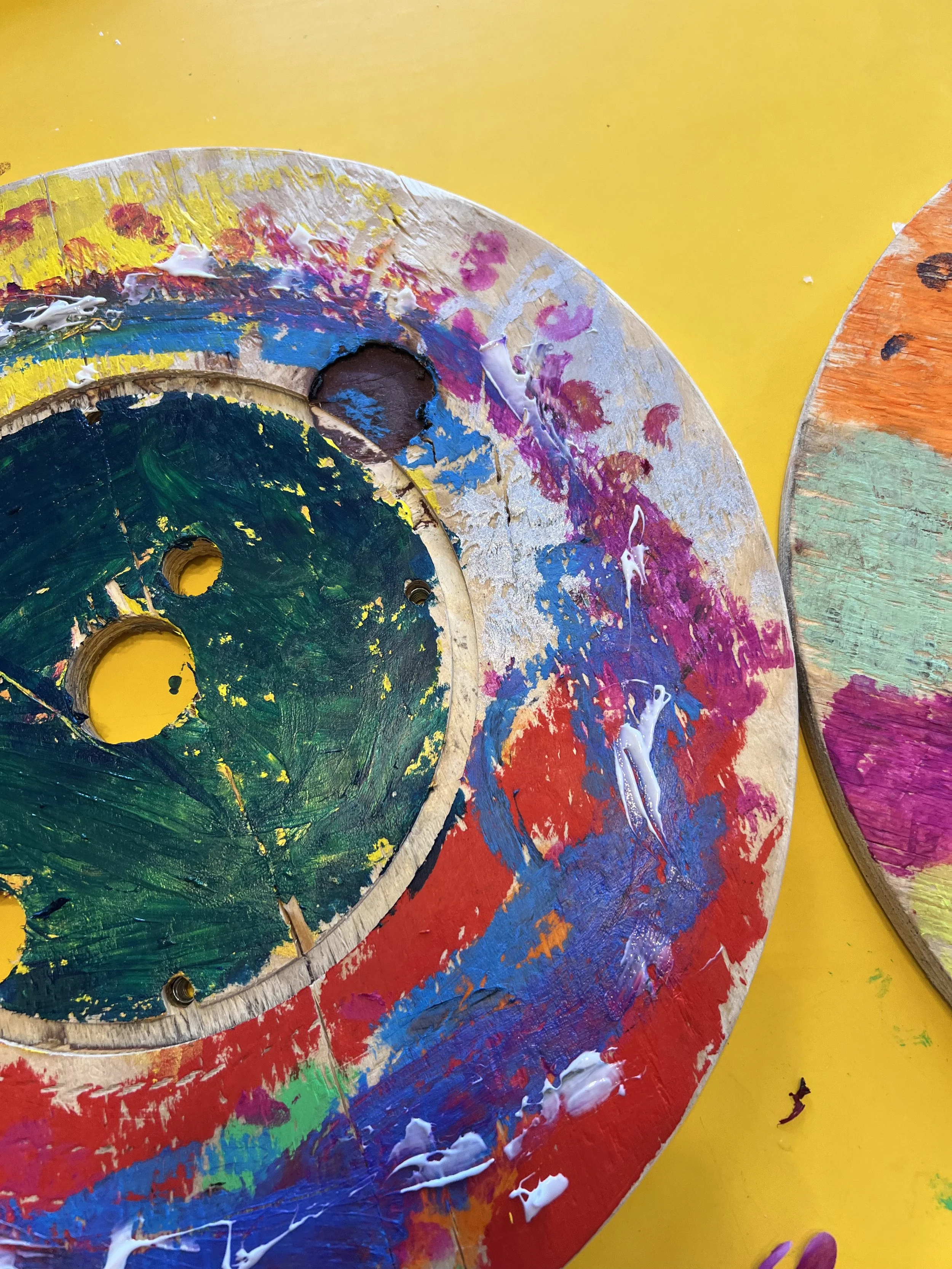 Painted wooden circular objects with multiple colors and splatters of white paint, placed on a yellow surface.