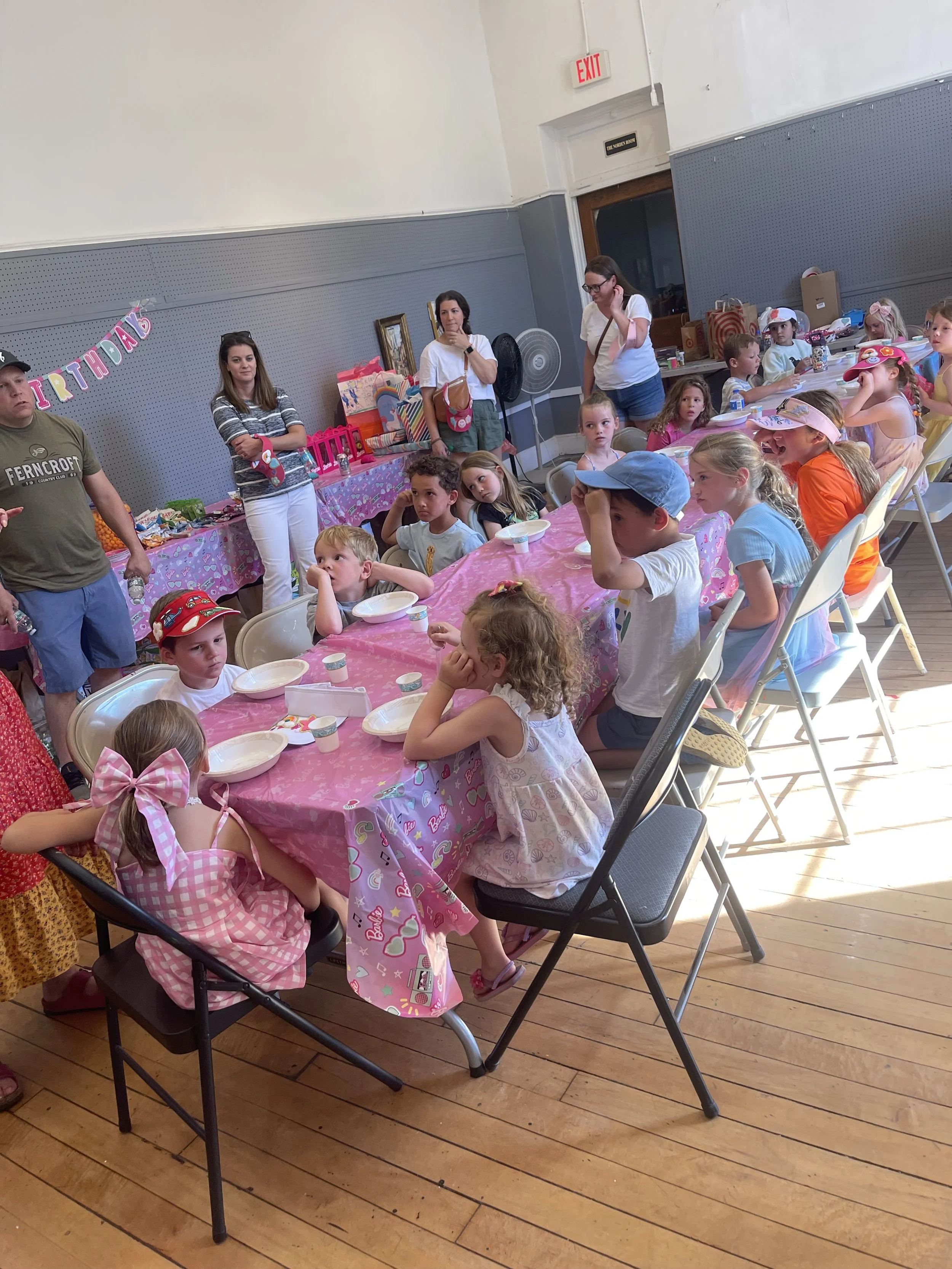 Children seated at a table with pink tablecloth, attending a birthday party with party hats and decorations, while adults stand nearby.