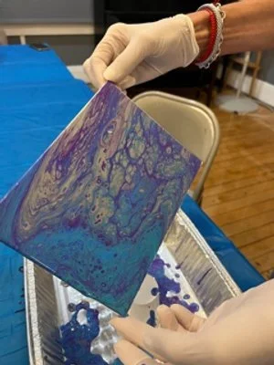 Close-up of a person wearing gloves holding a small square canvas with purple, blue, and teal marbled paint design.