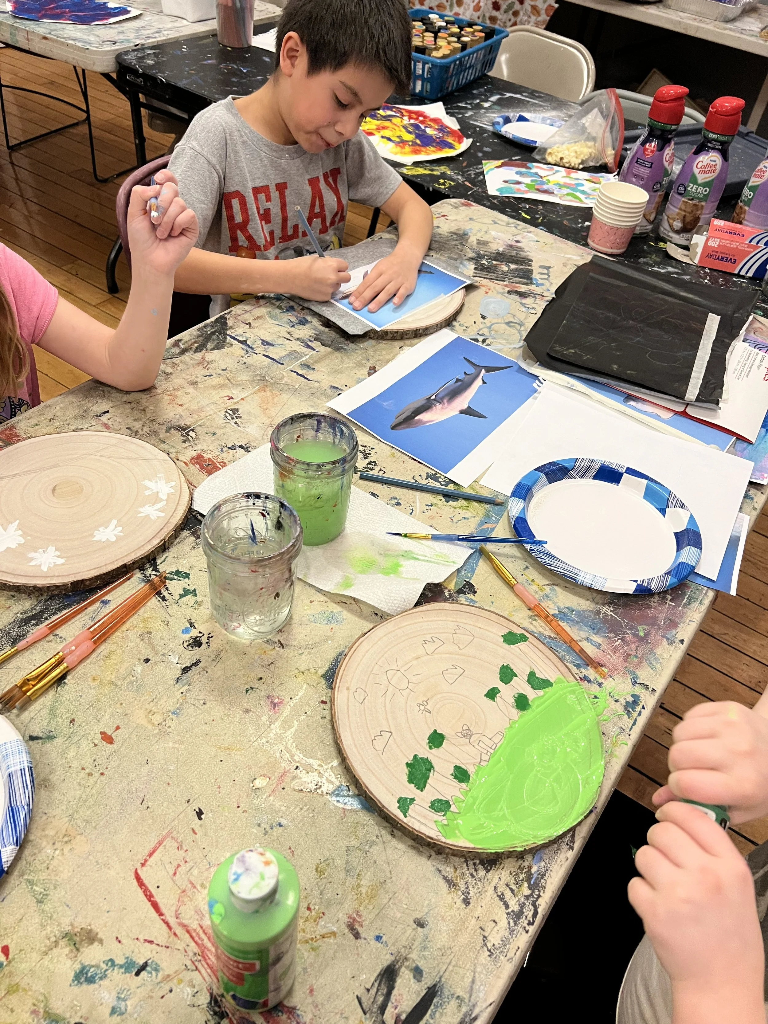 Children painting on circular wooden canvases surrounded by art supplies in an art studio.