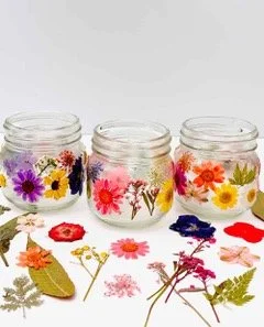 Three glass jars decorated with colorful paper flowers and leaves, arranged on a white surface with additional paper flowers and leaves spread out in front.