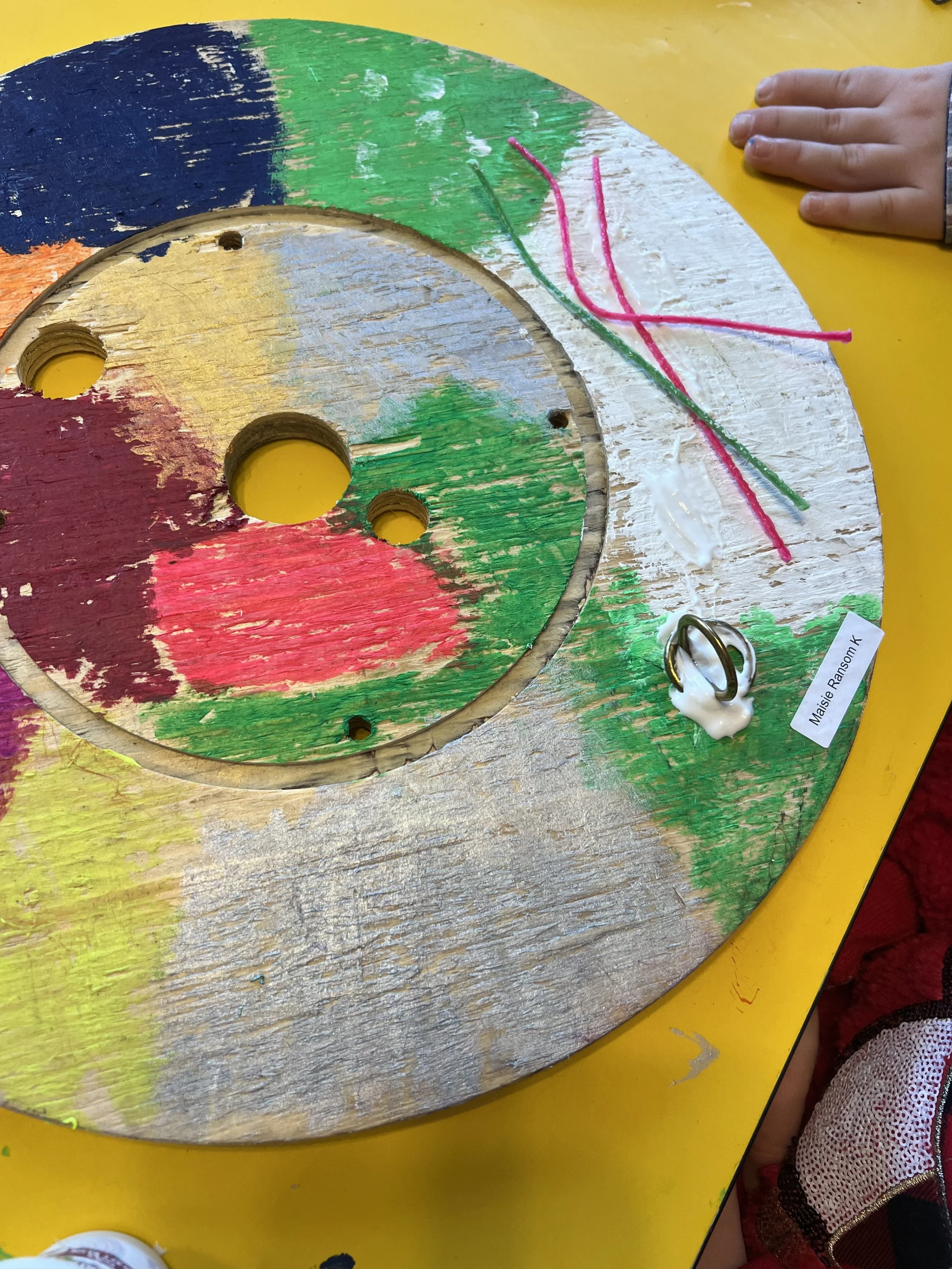 Painted wooden circular spinning wheel with colorful sections in green, yellow, blue, red, and white. There are three holes in the center and a handle attached to a white blob on the right side. Pink and green pipe cleaners are on the white section,