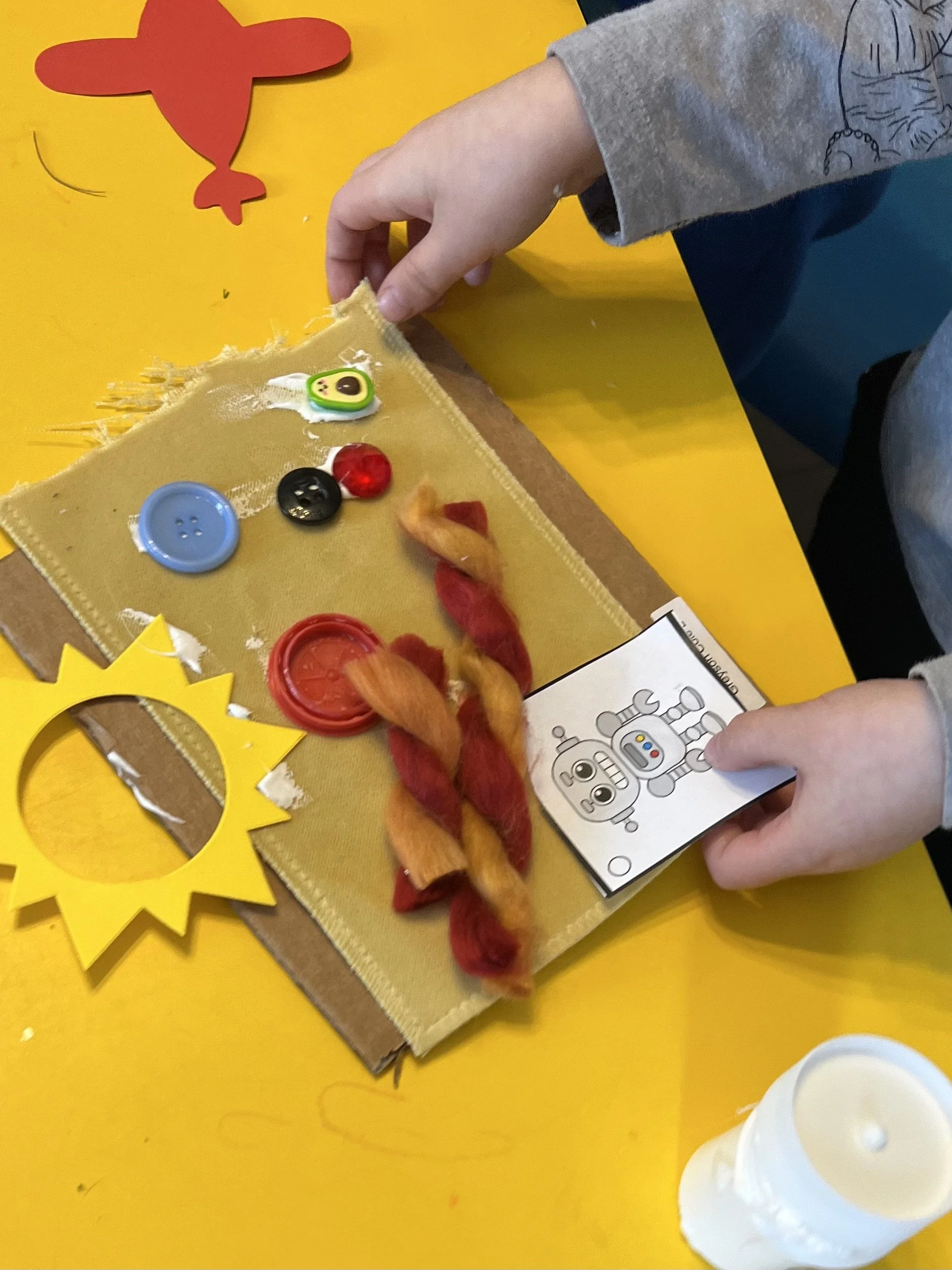 Child's hands assembling a craft project with colorful buttons, yarn, and a paper robot drawing on a yellow table with a red paper airplane and yellow paper cutout.