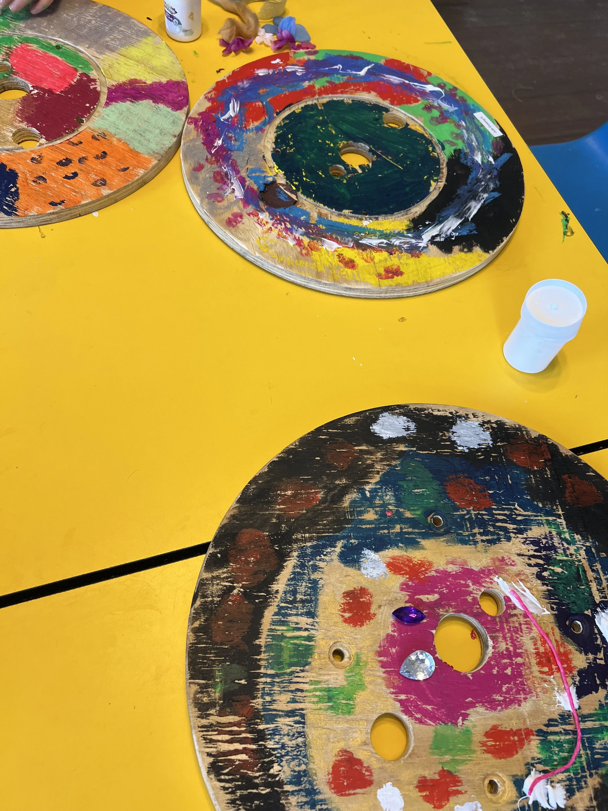 Colorful wooden discs with paint, glitter, and decorations on a yellow table.