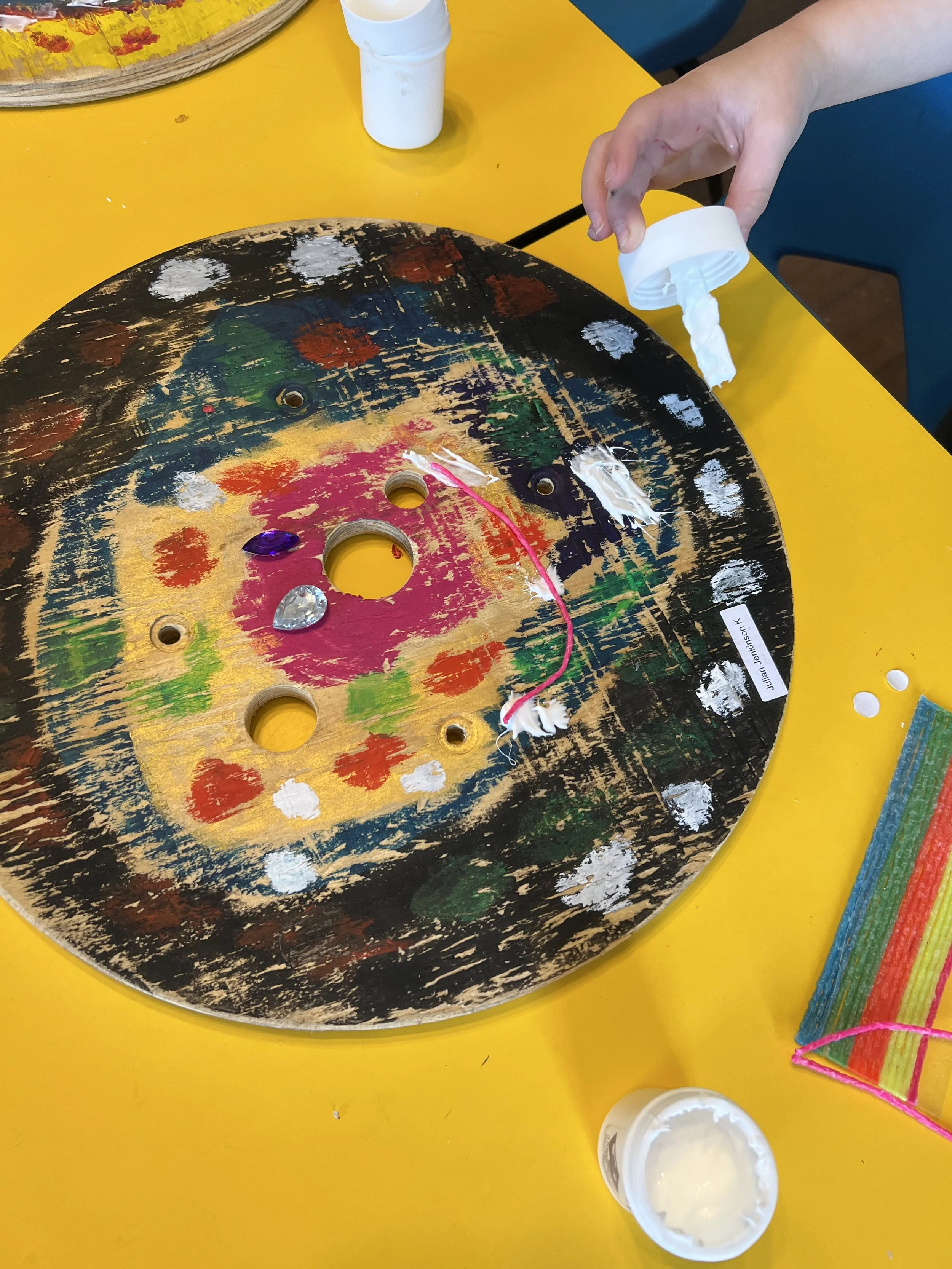 Colorful wooden round tapaz with white paint samples and a small container of white paint on a yellow table, and a hand pouring white paint from a container.