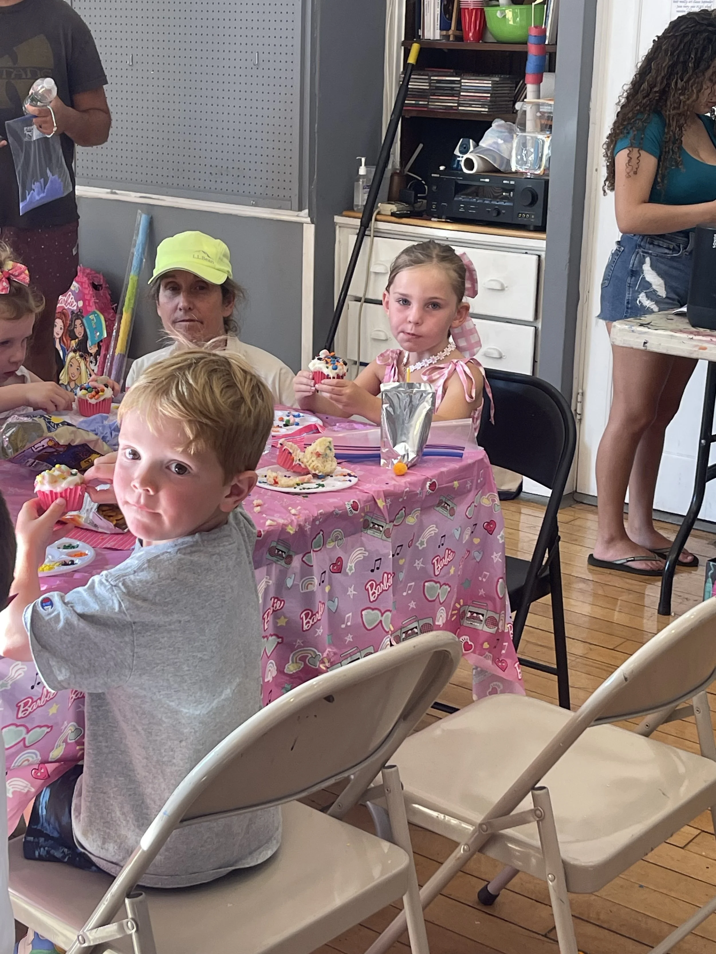 Children sitting at a birthday party table with pink Barbie-themed tablecloth, eating cupcakes with colorful sprinkles, in a room with wood flooring and shelves in the background.