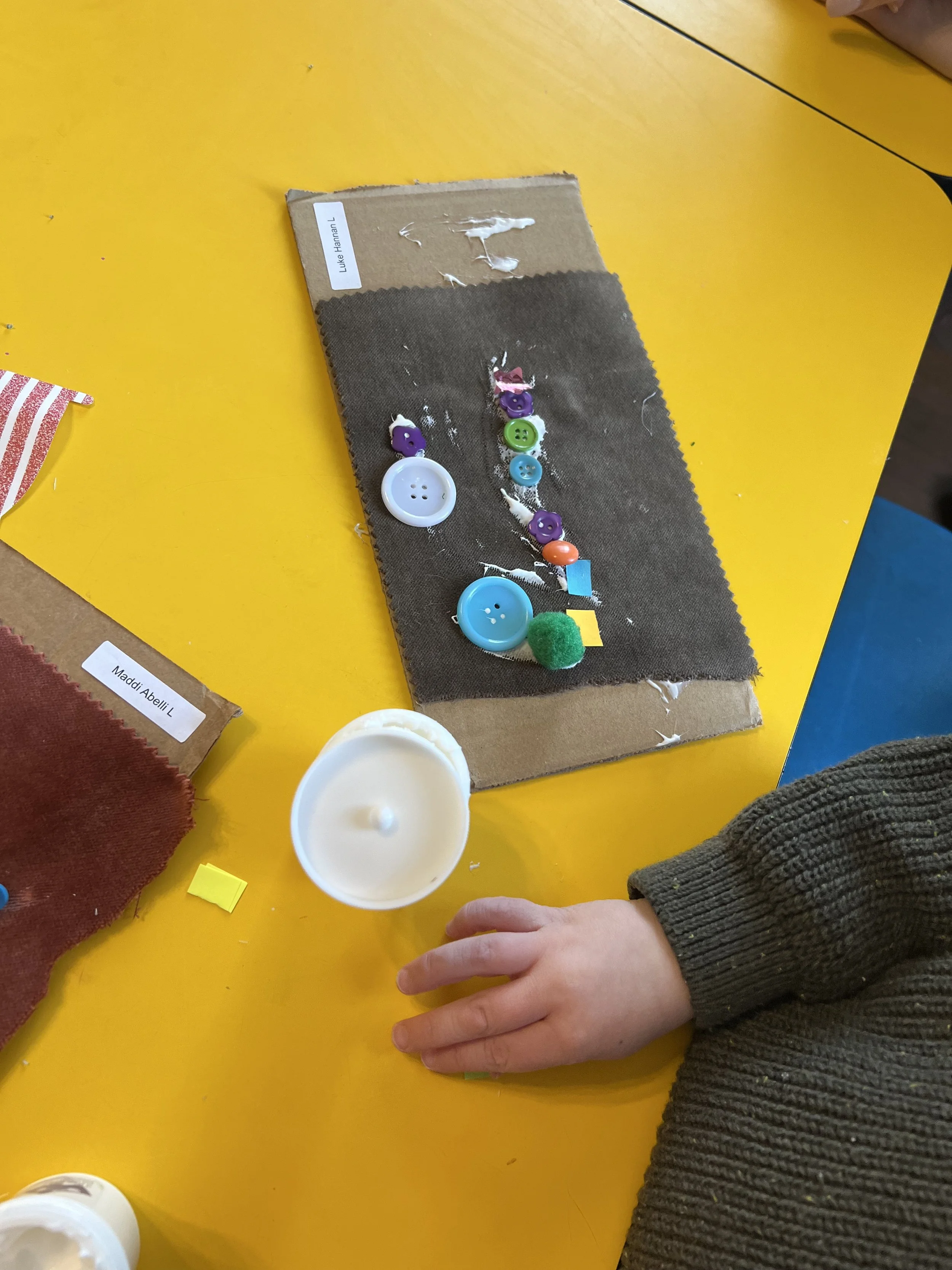 Child's hand resting on yellow table next to fabric swatches and a cardboard strip with various buttons and craft materials for a creative activity.