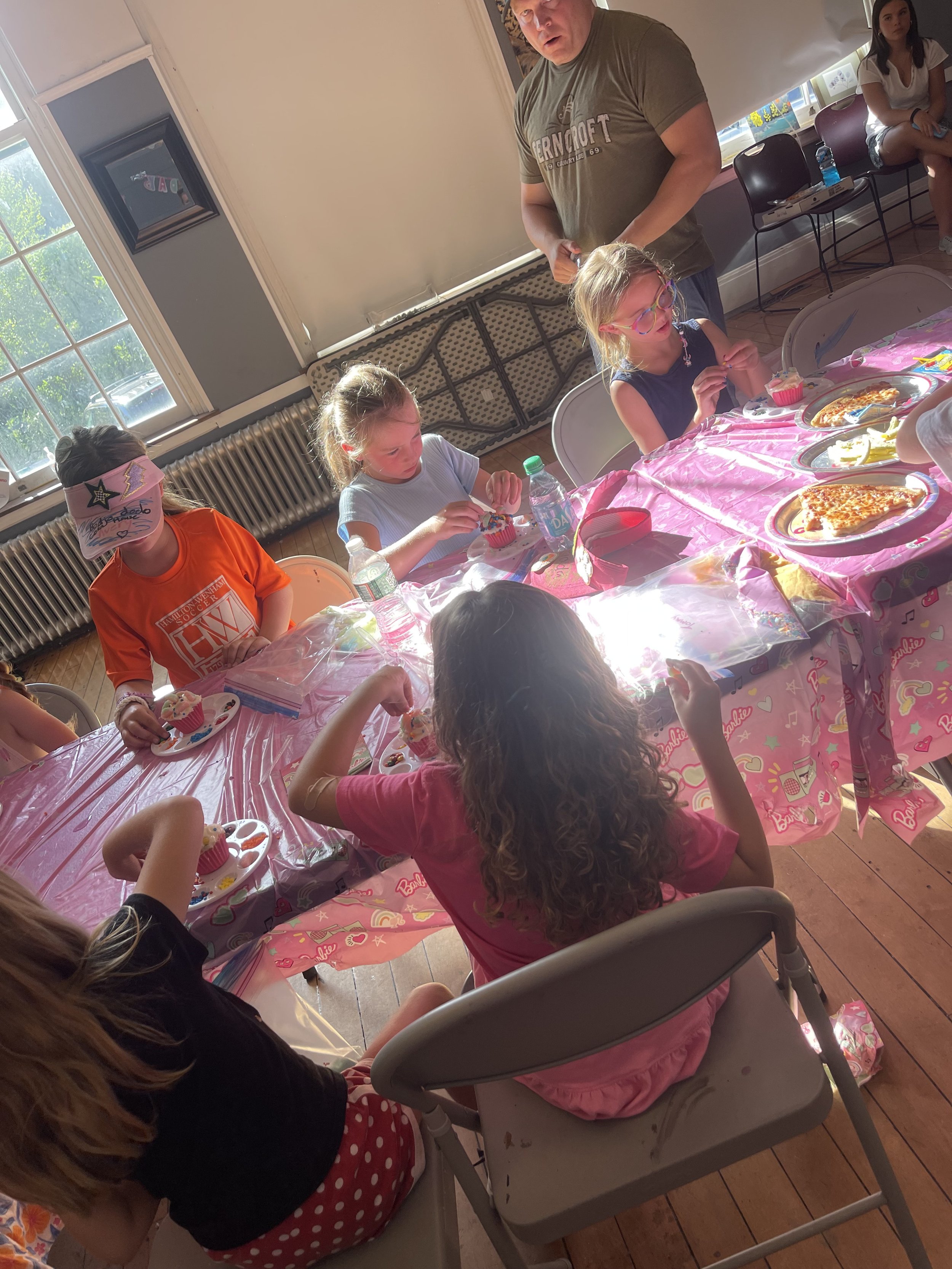 Children sitting around a table with pink Barbie-themed tablecloth, having a birthday party with cupcakes, pizza, and drinks. An adult man is standing nearby, and another girl is sitting against the wall.