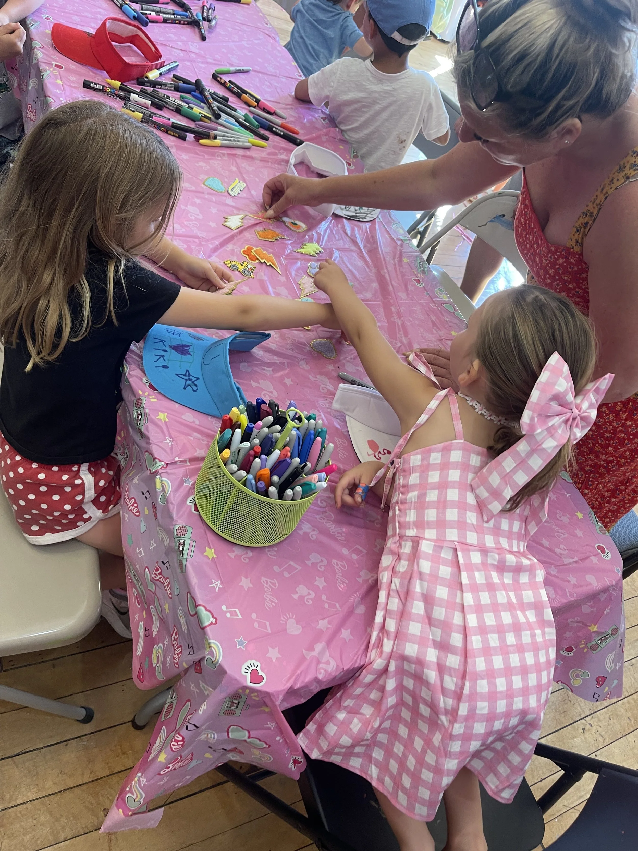 Children sitting at a pink tablecloth covered table, engaging in arts and crafts activity with colored pens, stickers, and pre-cut shapes. A woman is helping one girl place a sticker, and there are other kids in the background working on their projec