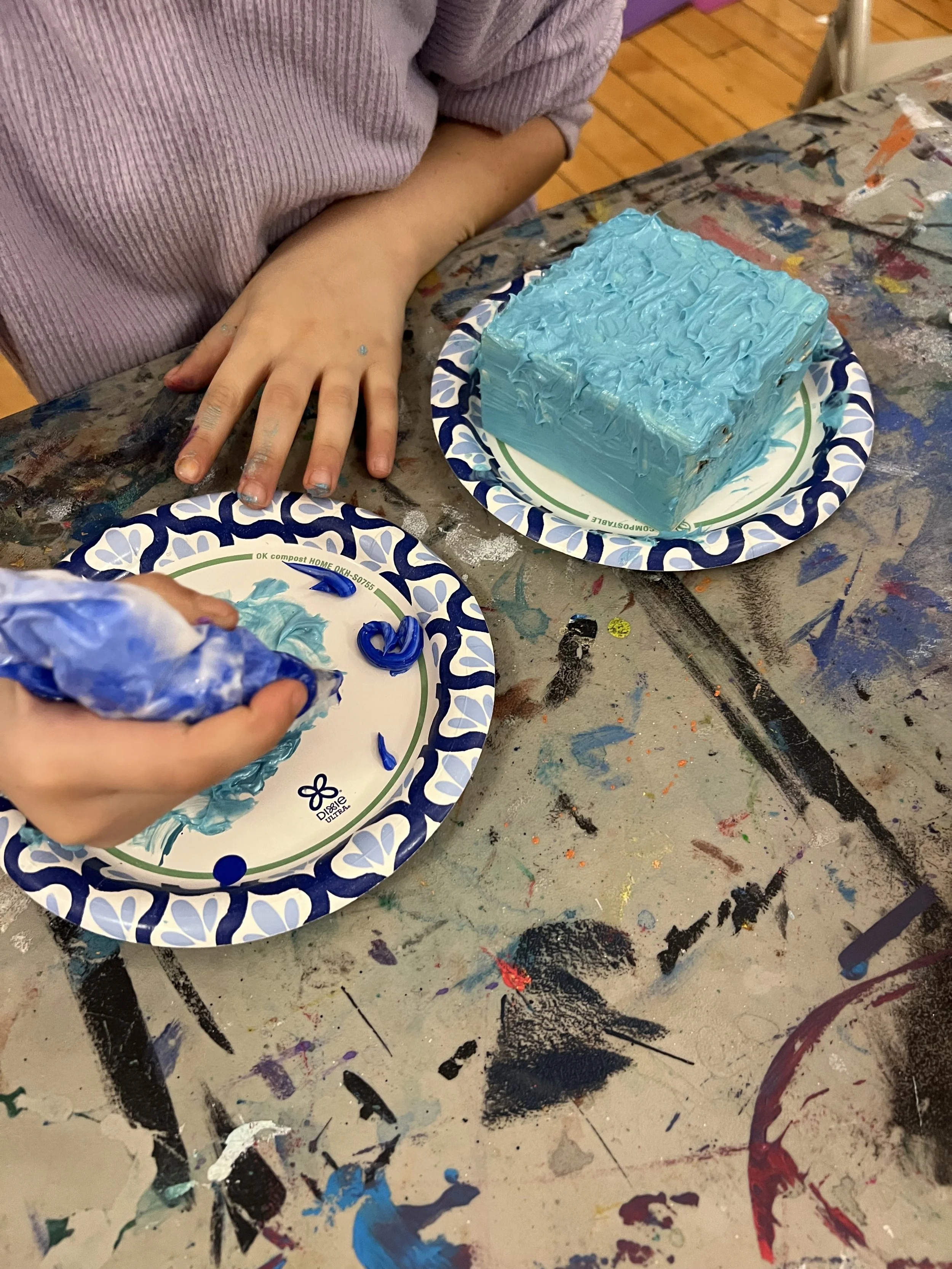 A child applies blue icing to a small round cake with blue icing. The cake is on a paper plate. The child's hand is visible, and the table has colorful paint stains and marks.