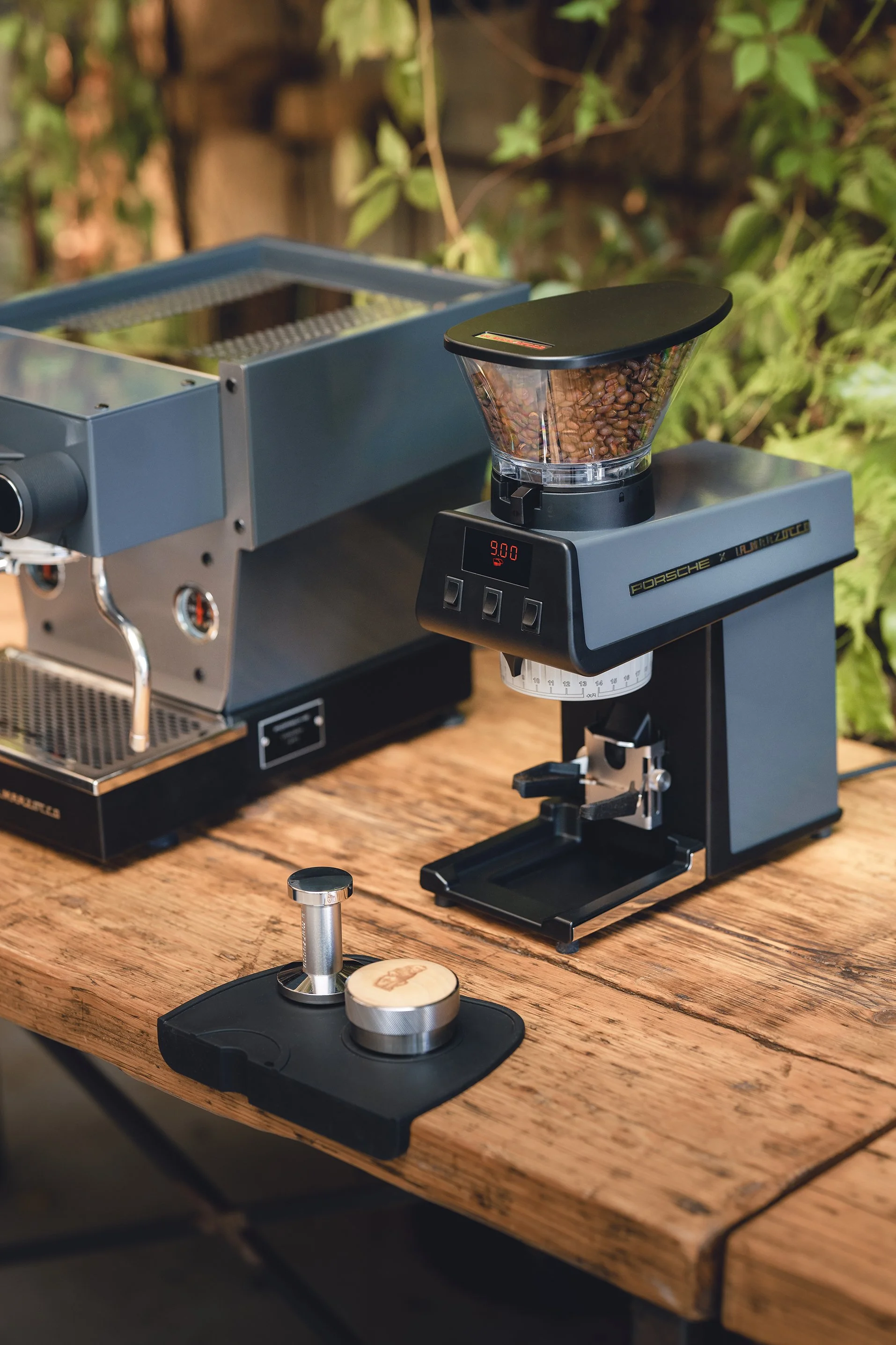 A coffee roasting machine, a coffee grinder, and a coffee tamper on a rustic wooden table. The coffee grinder contains roasted coffee beans.