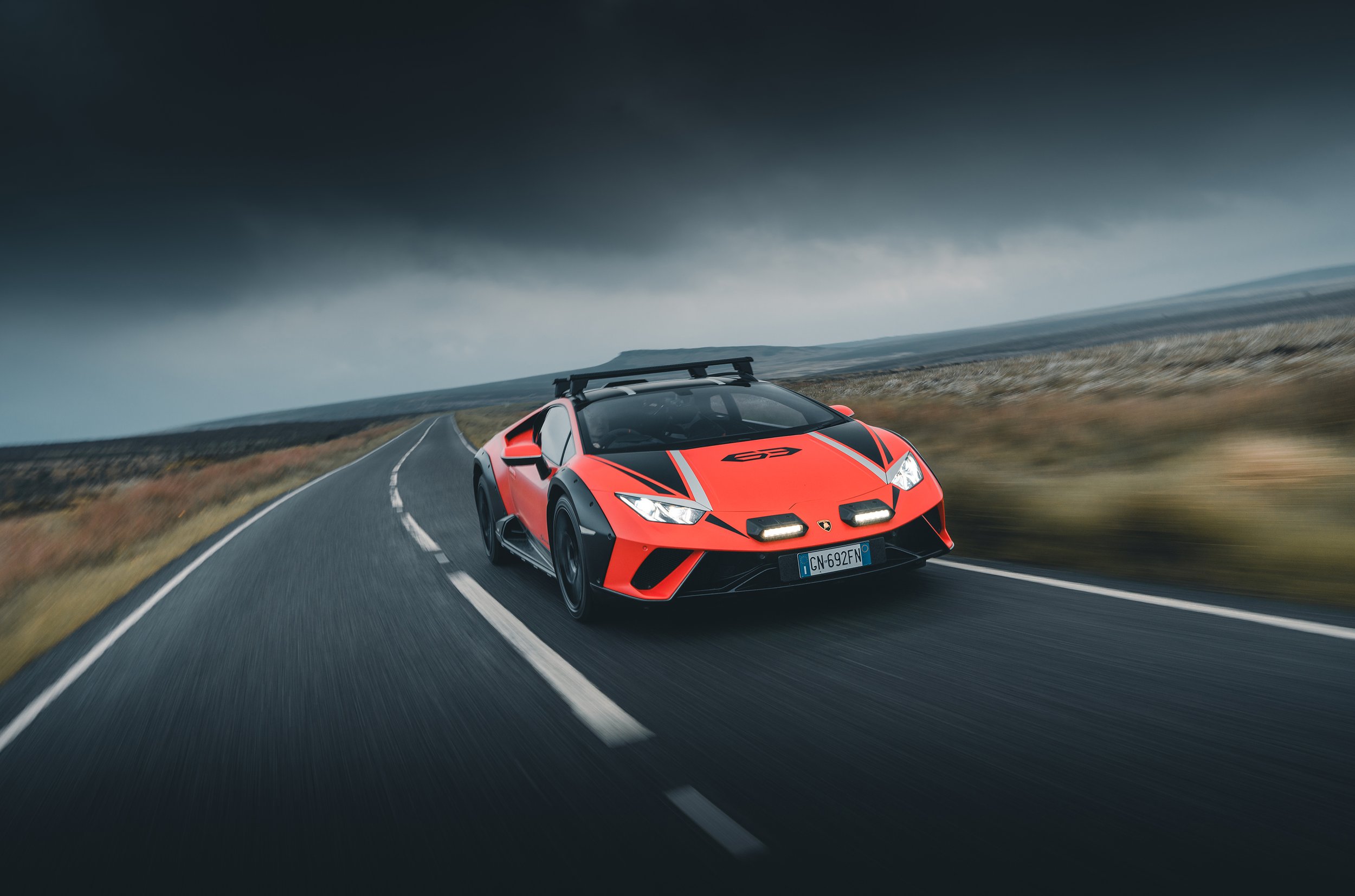 A red and black sports car driving on a winding road through a rugged landscape under a cloudy sky.