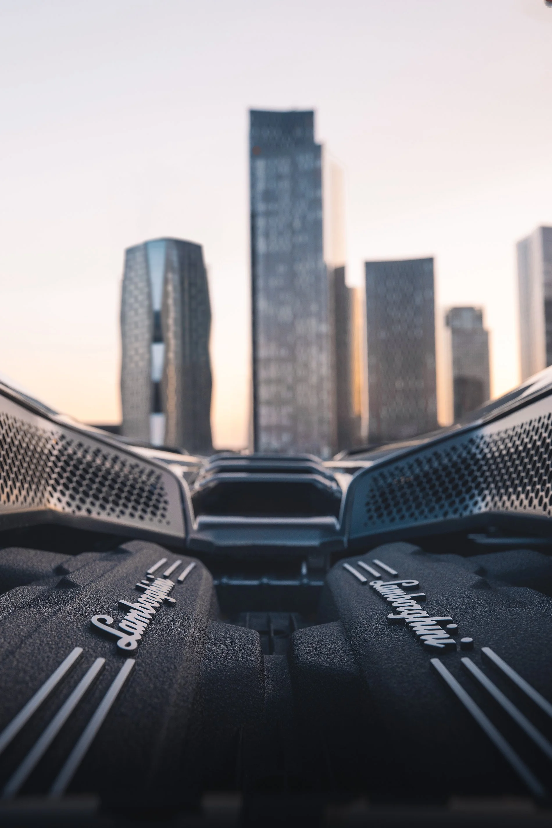 Close-up view of a Lamborghini engine with a city skyline in the background during sunset.