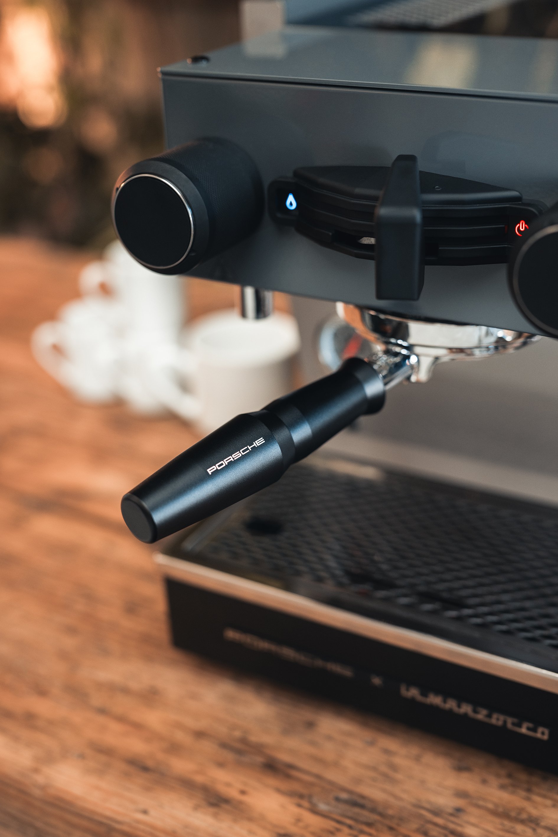 Close-up of a portafilter attached to an espresso machine, with the handle labeled 'Porsche,' on a wooden countertop.