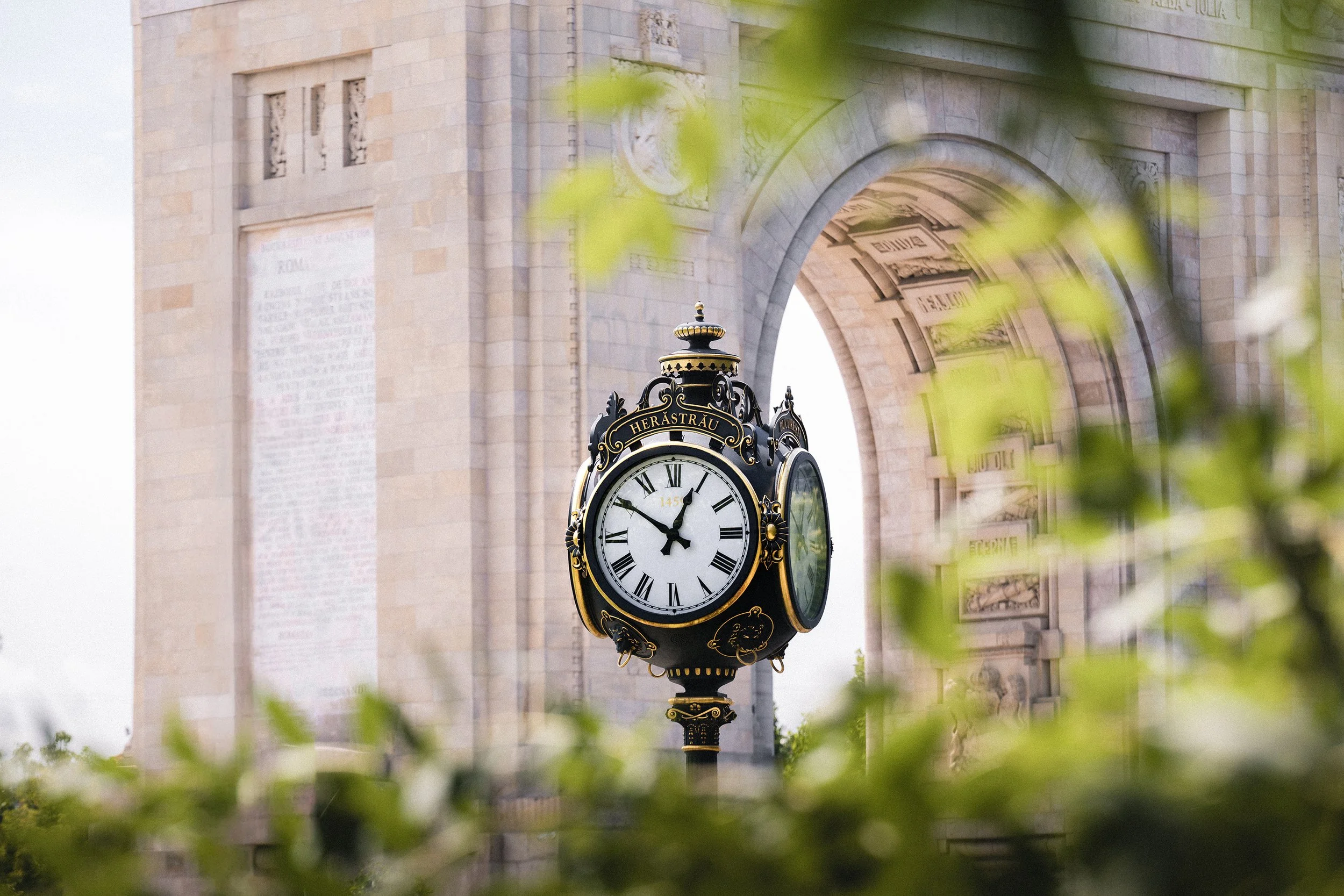 Historical clock near an archway in a cityscape, with greenery partially obscuring the view.