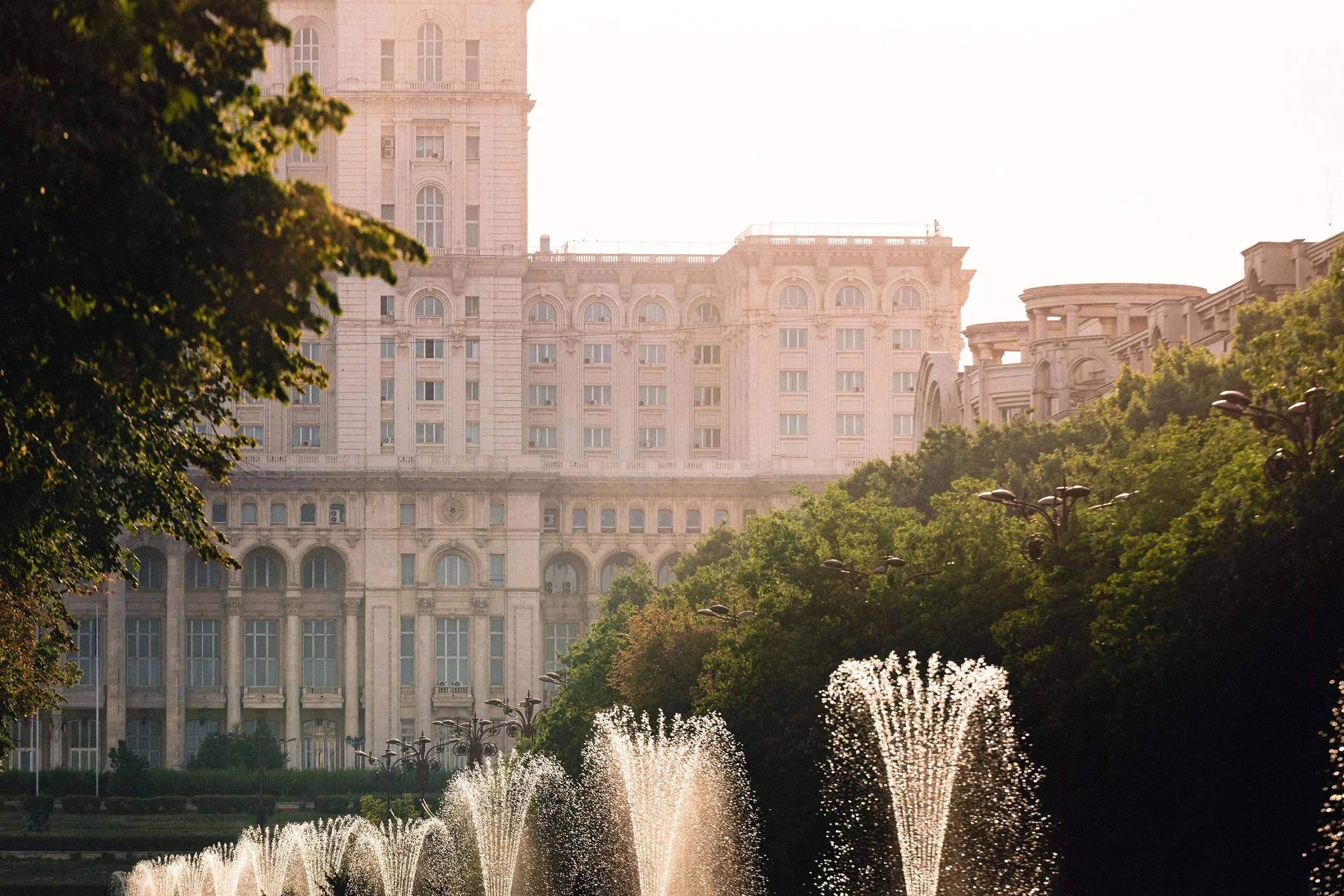 Large historic building with multiple windows, surrounded by trees, with fountains in the foreground in a park-like setting during daytime.